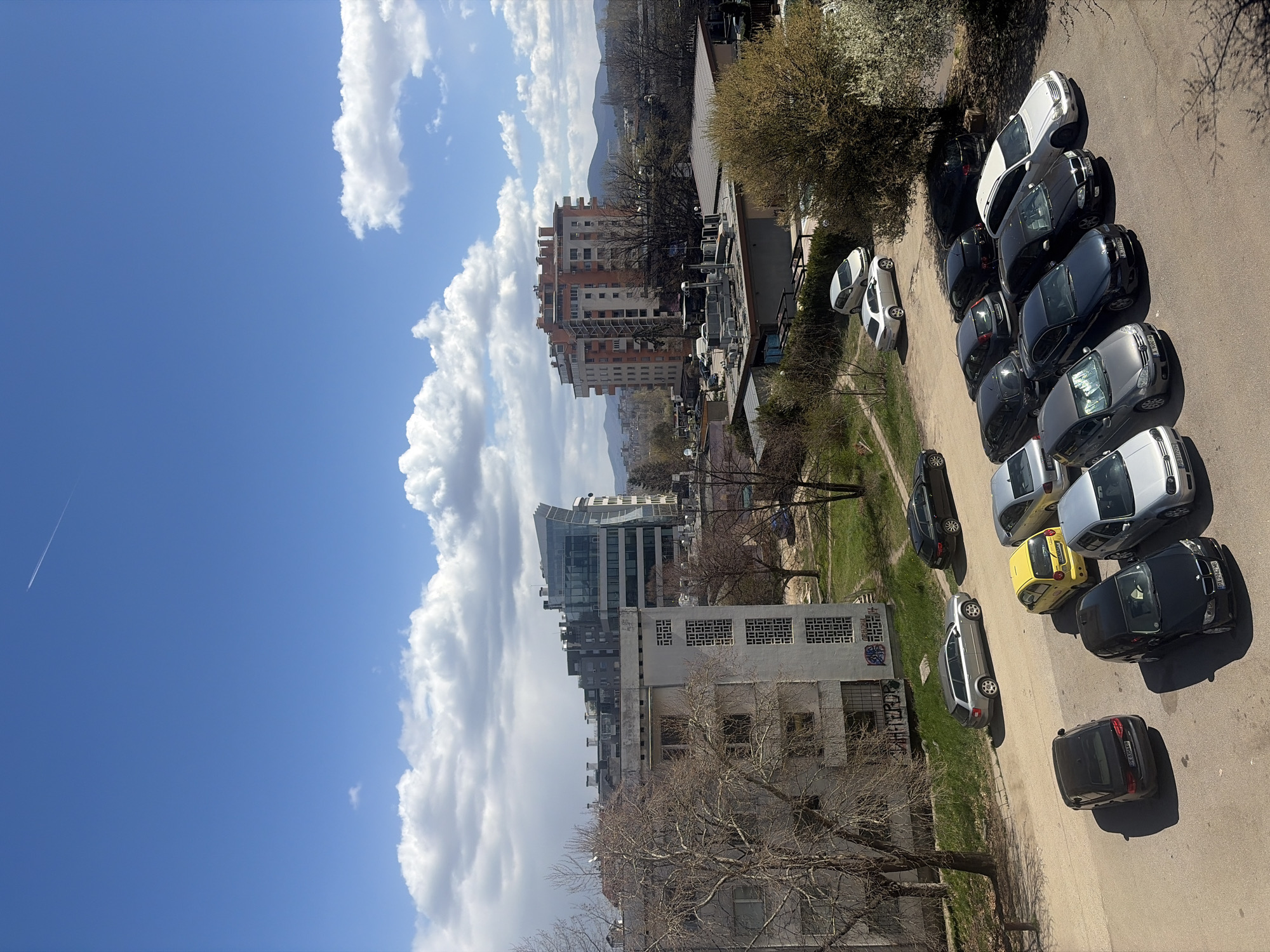 The image is a high-angle shot taken in Sofia, Bulgaria, capturing a mix of urban and natural elements under a bright blue sky dotted with fluffy white clouds. In the background, there are buildings of varying heights and architectural styles, including modern glass structures and older, possibly residential buildings. Further back, a mountain range is visible. A row of parked cars lines the right side of the frame along a paved street. Between the buildings and the street is a strip of greenery with trees in early spring, some still bare and others showing the beginnings of foliage. The overall impression is one of a sunny, early spring day in a residential area of Sofia.