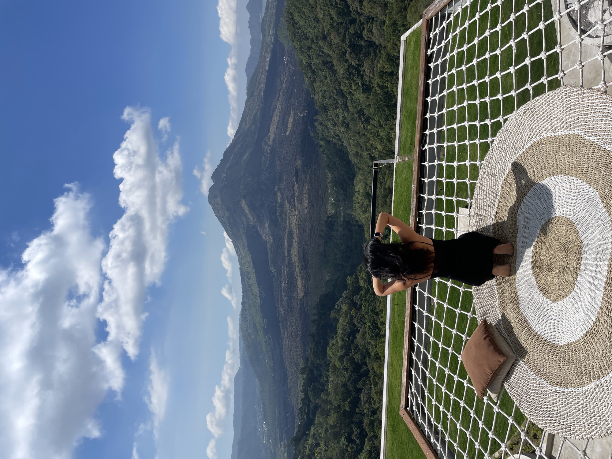 A woman with black hair, wearing a black dress, is kneeling on a woven rug on a net platform. She is facing away from the camera, with her hands behind her head, looking at a vast landscape. The landscape features a large, dark, volcanic mountain in the distance, with a lake visible to its right. The foreground and midground are covered in lush green forests. The sky is blue with scattered white clouds. The location is a scenic overlook in Kintamani, Indonesia, offering a view of Mount Batur and Lake Batur.