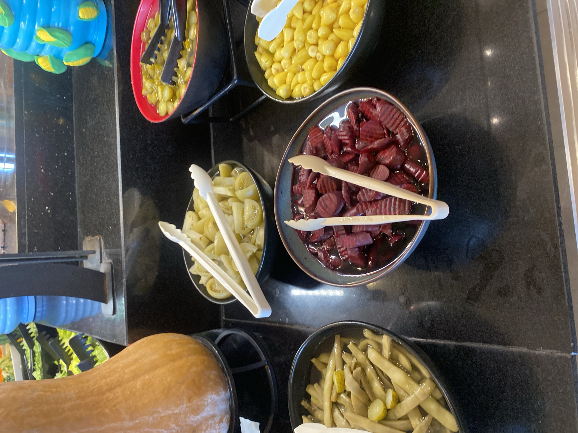 A close-up shot captures a food buffet display on a black countertop, featuring several bowls of pickled and preserved vegetables. To the left, a large, light orange butternut squash is prominently displayed, partially obscuring the view of a salad bar with green lettuce and black serving tongs behind it. In the center and right foreground, three bowls are visible. The leftmost bowl contains green beans and slices of pickles in brine. Adjacent to it, a bowl holds sliced pickled beets in a dark liquid. To the right, a bowl is filled with yellow pickled corn kernels. Black tongs are present in the bowls of green beans, beets, and corn, and white tongs are in the bowl of sliced tomatoes or similar light-colored pickled vegetables to the right of the squash. The background shows a blue dispenser with a ribbed texture, and a part of a decorative blue object with yellow corn cob details. The setting appears to be a restaurant or buffet. This scene is not specifically located in Aksu, Turkey; the context of Aksu, Turkey is not present in the visual information provided.