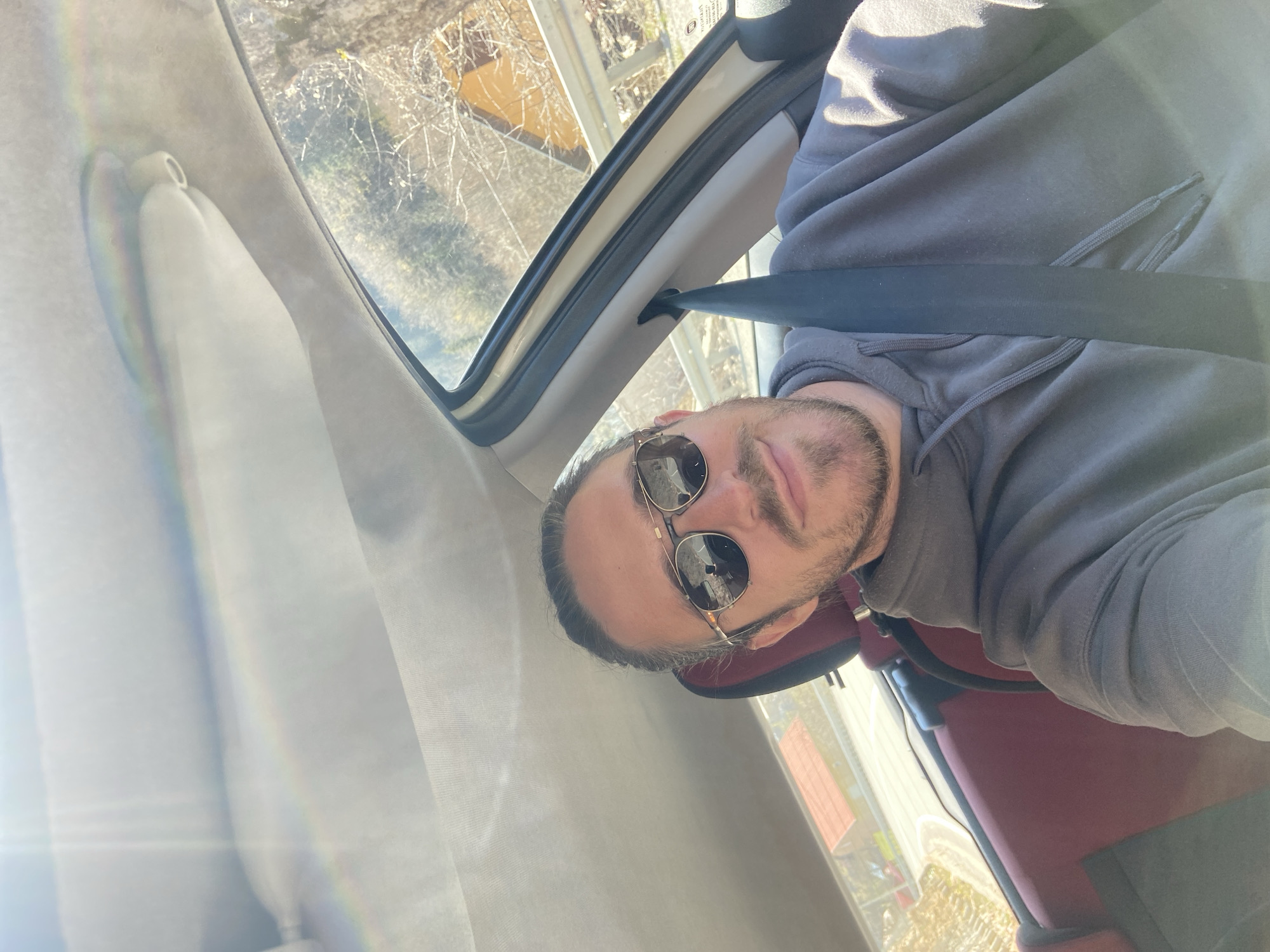 A man wearing sunglasses and a grey hooded sweatshirt sits in the driver's seat of a car. He is wearing a seatbelt. Outside the car window, trees and a building can be seen. The location is Lubnice, Montenegro.
