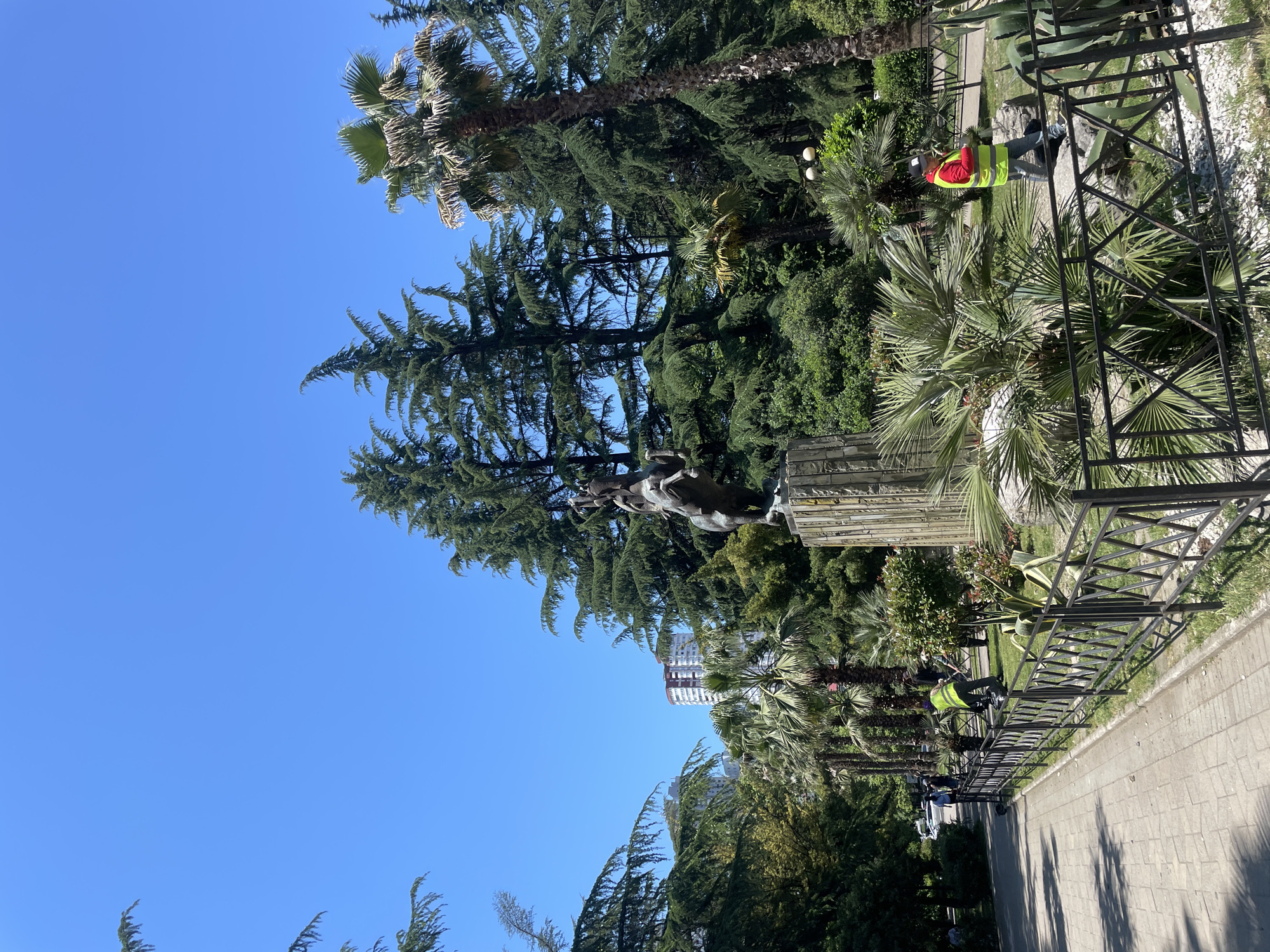 The image depicts a scene in Sochi, Russia, likely in a park or public square. A bronze equestrian statue stands prominently on a stone pedestal, surrounded by lush greenery including tall coniferous trees and palm trees. In the foreground, a paved walkway runs alongside a black metal fence. Several individuals are visible, some in yellow safety vests, tending to the park grounds. One person is actively working with what appears to be gardening equipment near the fence. In the background, a tall, modern building with a rounded top can be seen above the trees, indicating an urban setting. The sky is clear and blue, suggesting a sunny day.