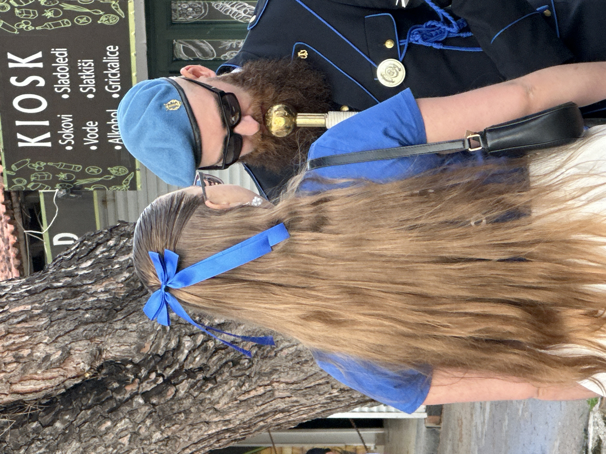 A medium shot captures a man in a dark blue uniform and a woman with long hair, both wearing blue shirts. The man has a full beard, sunglasses, and a light blue beret. He appears to be interacting with a golden scepter-like object. The woman's hair is light brown, tied with a blue ribbon. A black leather purse hangs over her shoulder.
In the background, a sign reads "KIOSK" with a list of items in Croatian, including "Sokovi," "Sokovi," "Vode," and "Grickalice." The presence of the Croatian language and the style of the kiosk suggest the image was taken in Montenegro, likely in the city of Budva. The setting includes a tree with rough bark.