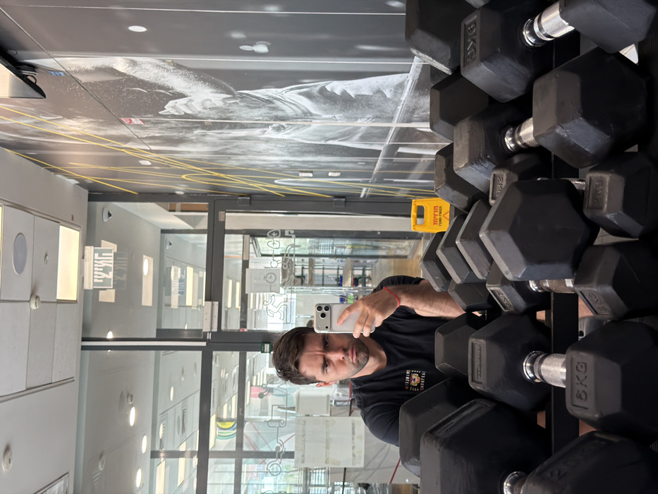 The image displays a mirror selfie taken by a male individual within a fitness facility in Tel-Aviv, Israel. The man, possessing short dark hair and facial stubble, wears a black t-shirt and a red string bracelet on his left wrist. He is holding a light-colored smartphone in his right hand, positioned to capture his reflection. His facial expression is neutral to slightly serious, characterized by furrowed brows and downturned lips.
In the immediate foreground, a rack of black, hexagonal dumbbells with chrome handles is prominent, partially obscuring the lower half of the man's body. Specific weights such as 8KG and 10KG are visible on some dumbbells.
The reflected background reveals features typical of a gym. To the right, a large black and white mural depicting a weightlifter, accented with yellow diagonal lines, covers a wall. To the left, a glass partition and door lead to another interior space. Above the glass door, an emergency exit sign with Hebrew text ("יציאה") is visible. Through the glass, additional fitness equipment and bright interior lighting are discernible. On the floor to the right, a yellow A-frame "wet floor" sign, also with Hebrew text ("זהירות רצפה רטובה"), is present. The ceiling features suspended acoustic tiles, recessed circular lights, and a rectangular light panel.