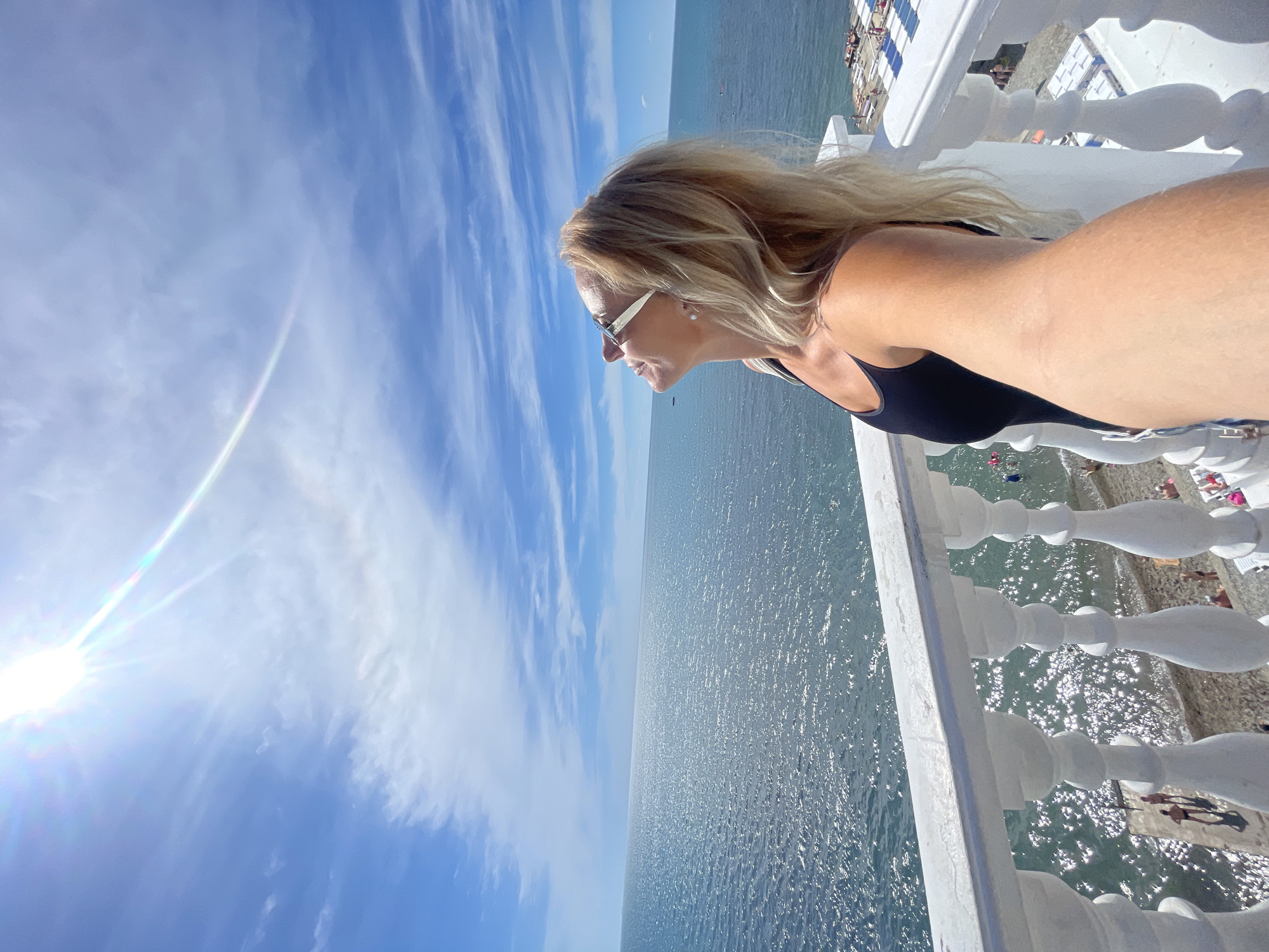 This media file features a woman in a black swimsuit and sunglasses standing on a balcony overlooking the sea. The sun is bright, indicating daytime, and the sky is blue with wispy clouds. The sea is sparkling with sunlight, suggesting a clear and sunny day. In the background, a beach with sunbathers and umbrellas is visible, along with the distant horizon. The balcony has a white ornate railing. The image appears to be a selfie, taken from a slightly high angle, with the woman looking out towards the sea. The scene suggests a relaxed vacation or leisure activity at a seaside resort. There is no visible text in the image. The location is Anapa, Russia.