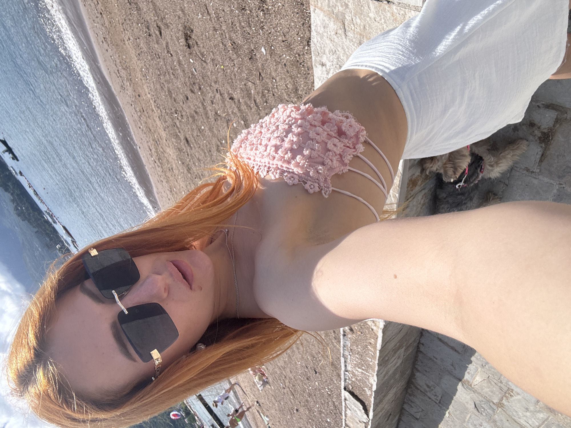 The image displays a young woman taking a selfie outdoors in Budva, Montenegro, under bright daylight. The photograph is rotated 90 degrees clockwise.
The primary subject is a woman with long, reddish-blonde hair. She wears large, black square sunglasses with distinctive gold accents on the frames. Her mouth is slightly open, and she has a delicate silver chain necklace around her neck. She is dressed in a light pink crop top with an intricate, textured design, possibly floral or lace, and thin straps that wrap around her midsection. Below the top, she wears a loose, white skirt.
She appears to be sitting or leaning on a stone structure, which could be steps or a low wall. In the background, a calm blue body of water, likely the sea or a large lake, reflects sunlight, creating shimmering patterns. Alongside the water is a sandy beach, where distant figures of other people are faintly visible. Small, dark shapes on the water in the distance might be boats. Partially obscured beneath her white skirt, a small, fluffy dog is visible, possibly lying down, with a hint of a red collar or leash.
The setting suggests a sunny, clear day at a coastal location, emphasizing leisure and outdoor activity. There is no visible text in the image.