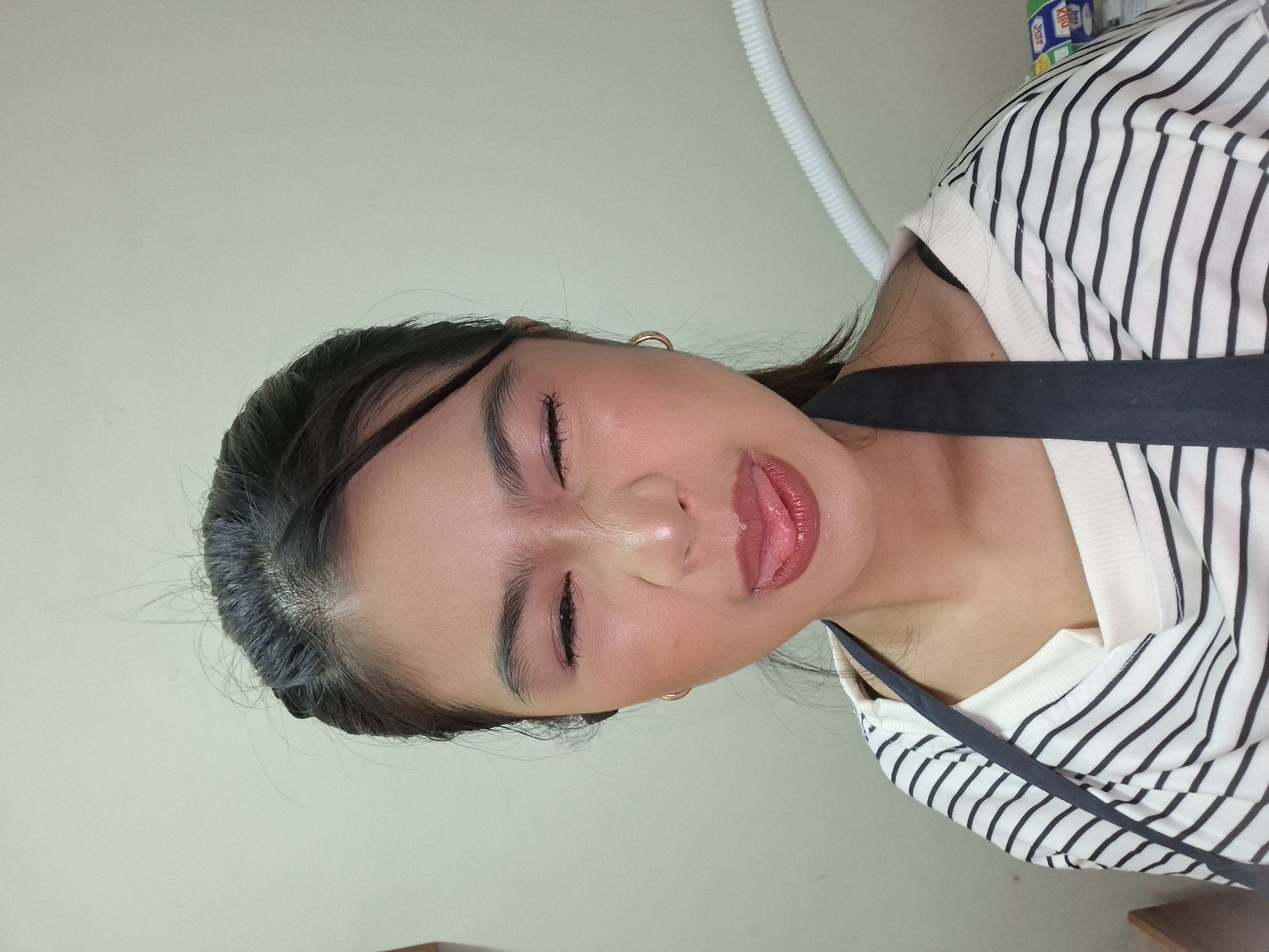 A young woman with dark hair pulled back is making a silly face, eyes squeezed shut and tongue sticking out, in a close-up shot. She is wearing a white and black striped shirt with a dark apron over it. The background is a plain, light-colored wall, and a white tube is visible on the right side of the frame. There are no specific details in the image that suggest the location is Kuta, Indonesia.