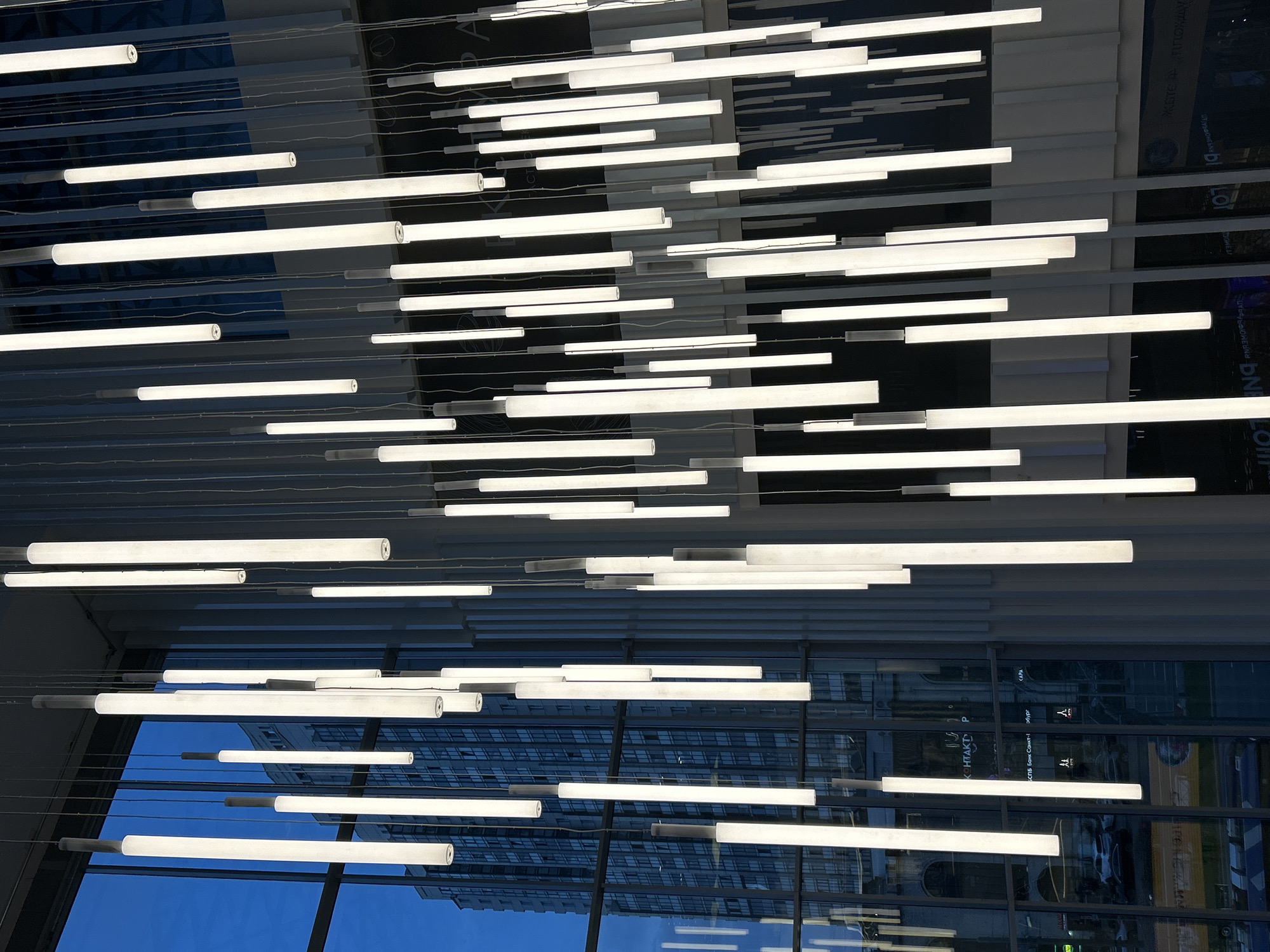 The image shows a modern lighting installation in Saint Petersburg, Russia. Numerous horizontal, linear LED light fixtures are suspended from the ceiling by thin cables. The background consists of a reflective glass facade, mirroring the surrounding urban landscape. The reflections include buildings, sky, and possibly people and vehicles, although these are difficult to discern clearly.