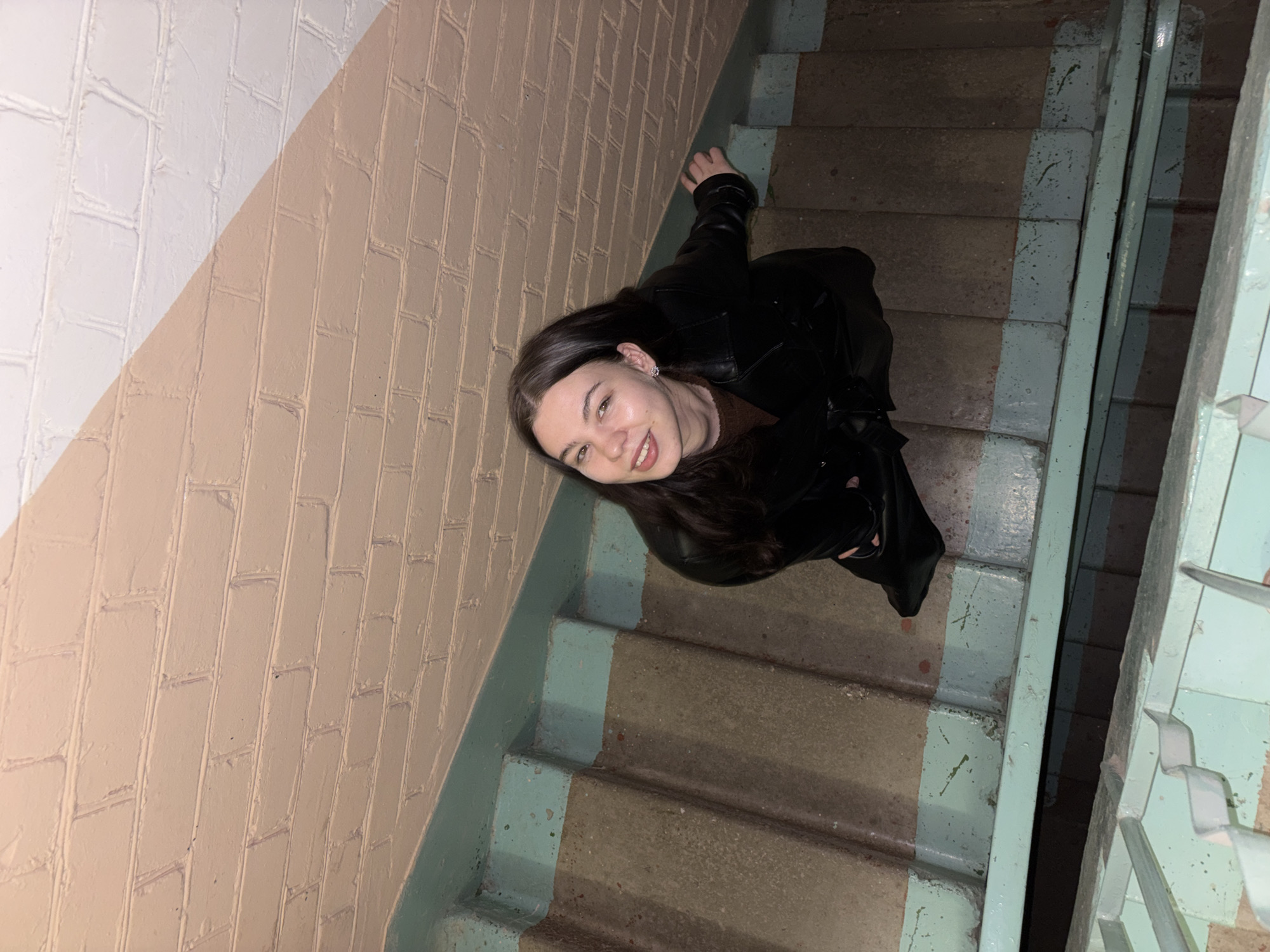 The image captures a young woman posing on an indoor concrete staircase, looking up towards the camera with a wide smile. She has long, dark hair and is dressed in a black leather jacket over a brown top, with silver stud earrings visible. The photograph is taken from an elevated perspective, looking down the stairwell.
The setting is a utilitarian stairwell, consistent with an apartment building or public access structure in Perm, Russia. The concrete steps are worn, and their edges and vertical risers are painted a faded, light blue-green. A metal railing, also painted in the same blue-green hue, runs alongside the staircase. The adjacent wall features a distinct two-toned painted brick pattern. The upper left section of the wall is white, while the larger lower section, extending parallel to the stairs, is a light beige or tan. The diagonal line separating these two colors creates a visual division on the wall.
The lighting in the stairwell is dim and artificial, indicating an interior space with little to no natural light, possibly suggesting it is nighttime or a perpetually low-light area. The paint on both the stairs and the railing shows signs of wear and chipping, suggesting the age and frequent use of the structure. The woman's expression conveys happiness and friendliness. There is no visible text in the image.