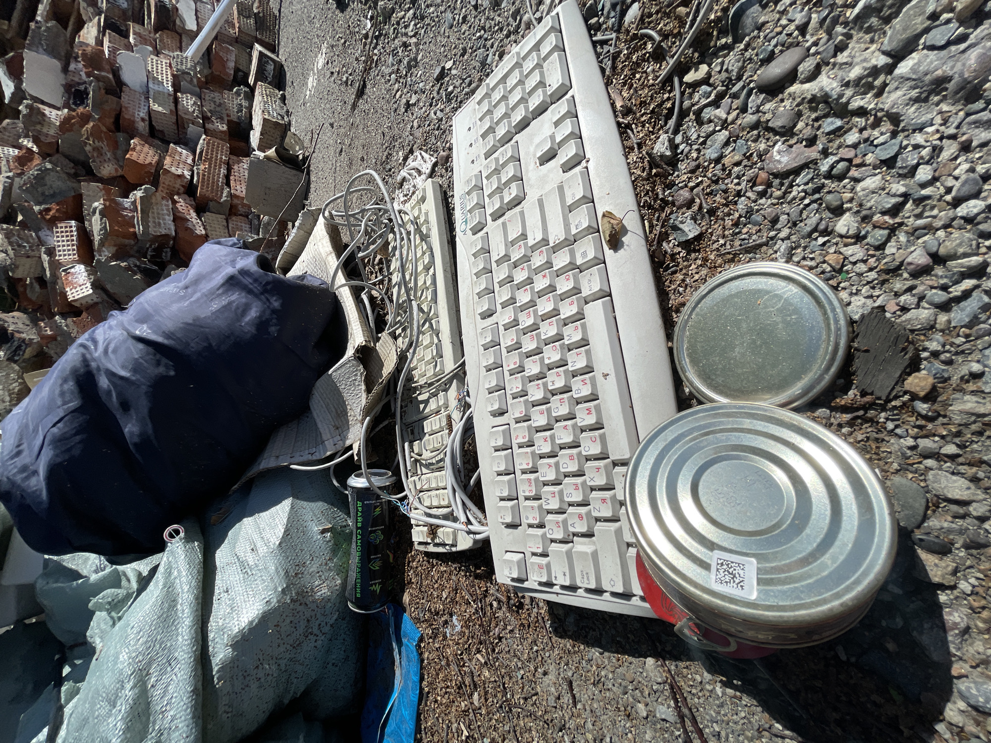 This image captures a close-up, high-angle view of discarded items on a gravelly and dusty ground, likely an outdoor waste area in Krasnoyarsk, Russia. The scene is dominated by two old, off-white computer keyboards, featuring both English and prominent Cyrillic (Russian) keycaps, with one bearing an "AQUARIUS" logo. Tangled cables and wires are visible around them.
To the left, a heap of broken bricks and concrete rubble forms a backdrop, suggesting a demolition or construction waste site. In the foreground and middle ground, a dark blue, crumpled fabric item, possibly a jacket or tarp, rests upon a larger, light green woven tarp. Adjacent to these fabrics is a black and green aerosol can with Cyrillic text, reading "ДРАЙВ САМОВЫРАЖЕНИЯ" (DRIVE OF SELF-EXPRESSION).
To the right of the keyboards, two round, metallic cans with smooth, dome-shaped lids are present; one has a small QR code sticker. The ground throughout is a mix of small stones, gravel, and dirt, with some dried leaves or debris scattered around. Bright sunlight casts distinct shadows, indicating a clear, sunny daytime. No people are visible, and the overall impression is one of abandonment and neglect, typical of a dumping ground or a site cleared of structures.