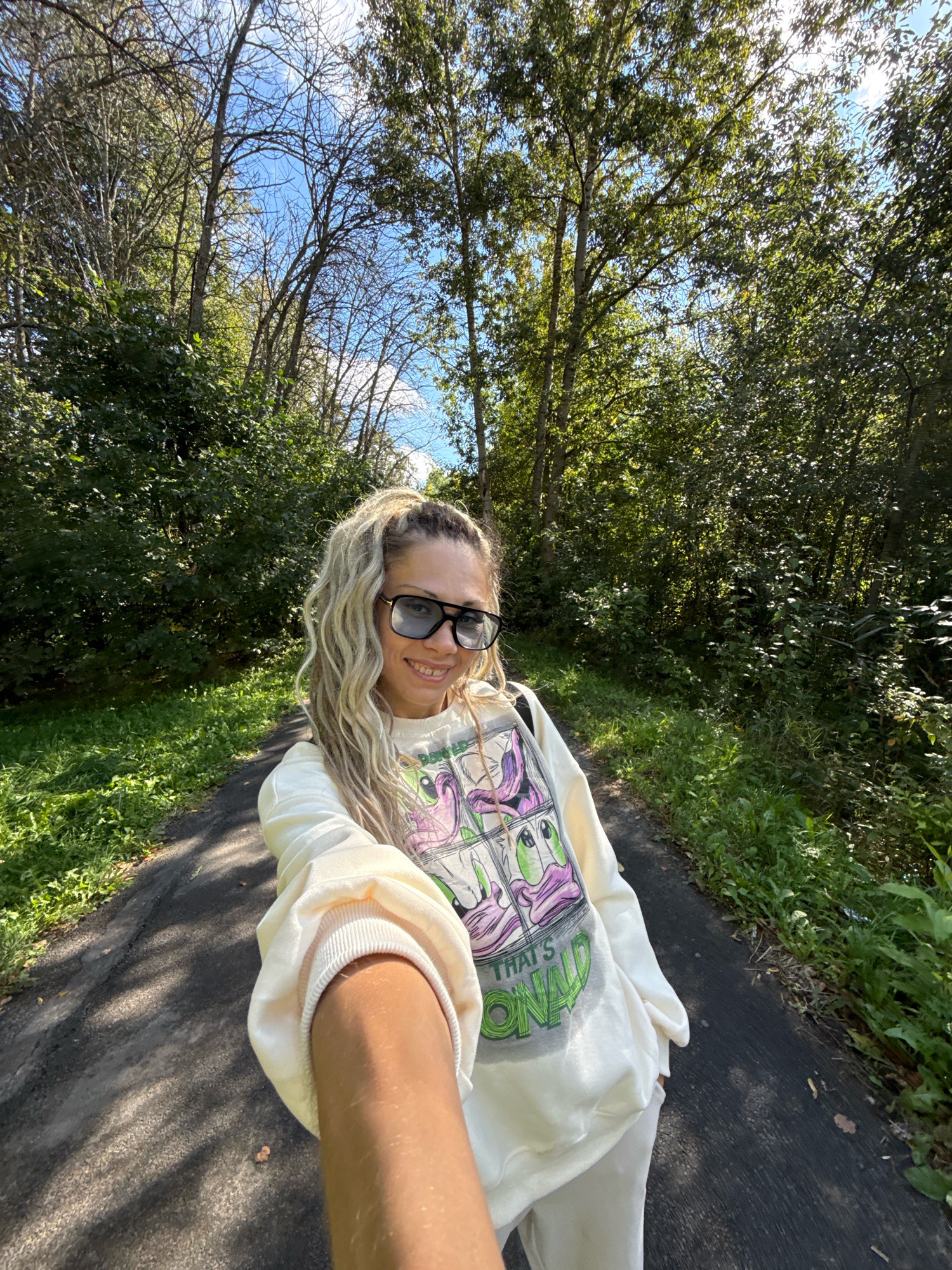 In the city of Rusanovo, Russia, on a sunny day with scattered clouds, a woman with blonde, wavy hair pulled into a high ponytail and wearing oversized black sunglasses is captured in a selfie. She is smiling and wearing a cream-colored sweatshirt with a cartoon graphic featuring green and purple characters, and the text "THAT'S ONALD." She is also wearing matching cream-colored sweatpants. The woman is standing on a paved path that winds through a wooded area. Lush green trees and foliage line both sides of the path, with sunlight dappling through the leaves, casting shadows on the ground. The overall scene suggests a pleasant outdoor activity, possibly a walk or jog, in a natural setting.