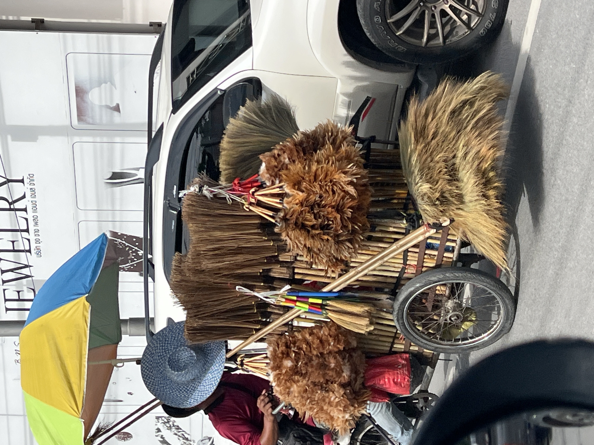 A person wearing a maroon shirt and blue jeans sits on a bicycle, holding a large blue hat. The bicycle is attached to a cart filled with brooms and feather dusters. A colorful umbrella shades the scene from above. In the background, a white SUV is parked next to a building with a sign that reads "JEWELRY". The location appears to be Baan Plai Laem, Thailand, based on the context provided. The street is paved and there are other vehicles in the foreground, suggesting a busy urban environment.