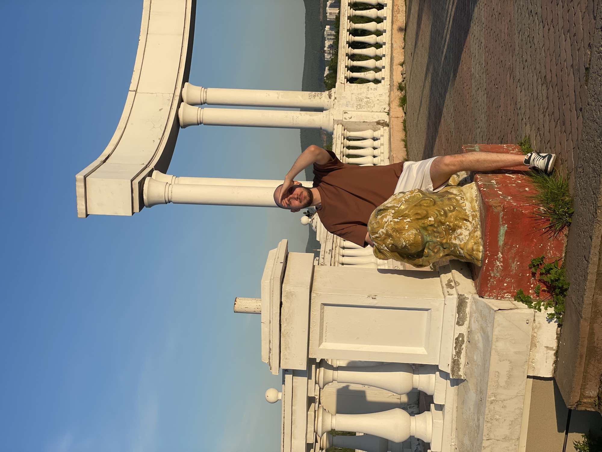 A man is captured lying down on his back on a weathered red and gold lion statue base, under a bright blue sky, in what appears to be a public architectural setting in Krasnoyarsk, Russia. The man, with a beard, wears a brown t-shirt, white shorts, and dark sneakers. His head is towards the center of the frame, and his legs extend to the right. His left hand is raised, shading his eyes as he looks up, likely at the sun or the camera.
The scene is dominated by a large, ornate white architectural structure to the man's left and above him. This consists of multiple classical-style pillars supporting a grand, curved archway that extends across the upper left of the image. A white balustrade-style railing runs behind the man, separating him from a view of distant, tree-covered hills dotted with buildings. Both the archway and the railing show signs of significant wear, with cracked paint, chipping, and faint, indecipherable graffiti marks on the balustrade.
The ground in the foreground and to the man's right is paved with reddish-brown bricks, illuminated by strong, warm sunlight that casts long shadows, suggesting either early morning or late afternoon. Patches of green grass and weeds are visible growing around the red pedestal base. The sky is uniformly clear and blue, with no clouds. No decipherable text is visible on any surfaces.