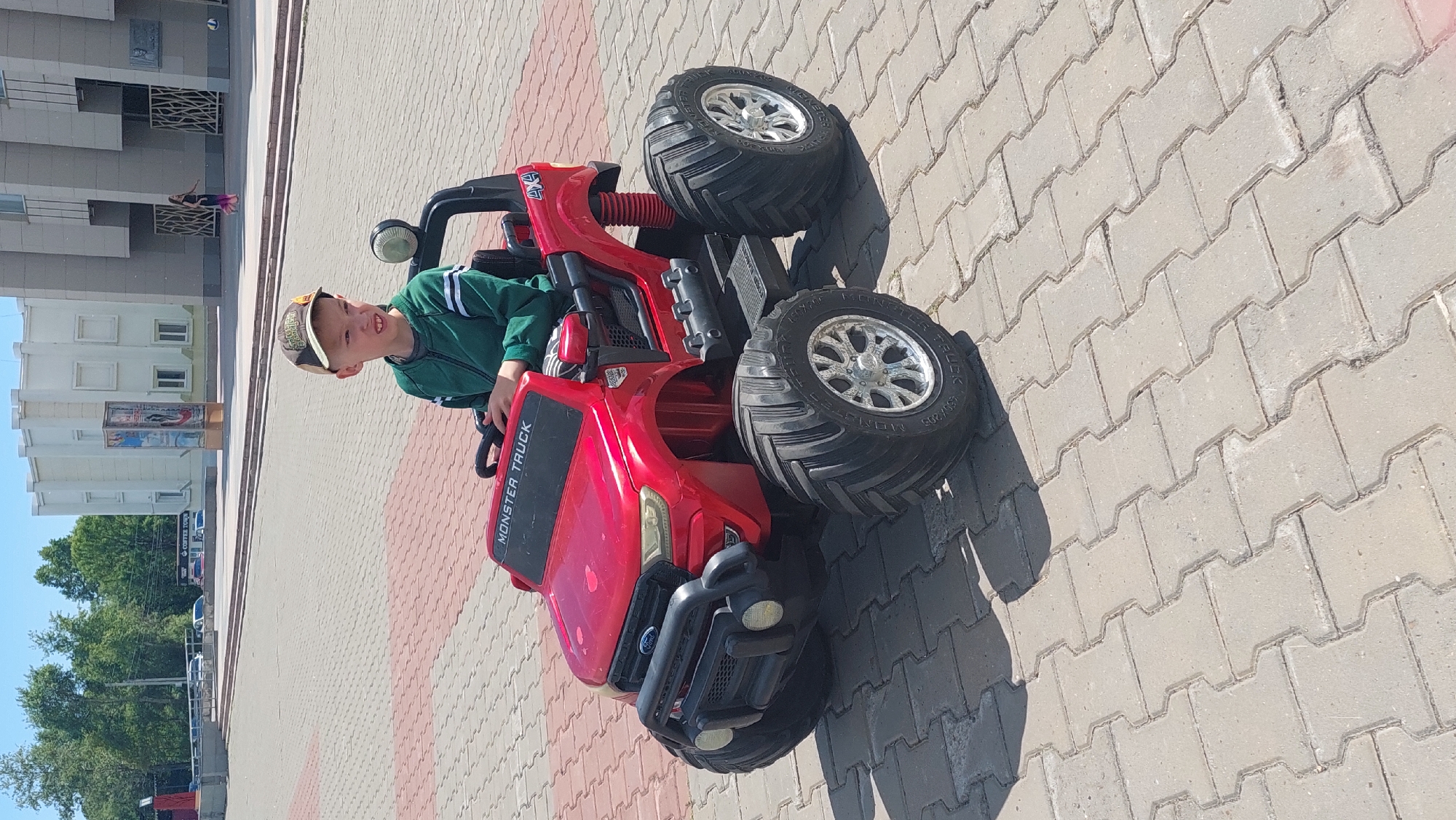 A young boy wearing a baseball cap and a green jacket is sitting in a bright red toy monster truck on a paved walkway. The truck has large, off-road tires and a black bumper with lights. The boy is looking forward with a slight smile. In the background, there are buildings, trees, and a blue sky, indicating it is daytime and likely sunny. The walkway is made of interlocking concrete pavers in shades of grey and pinkish-red. There are two people walking in the distance in front of a building. Visible text on the truck reads "MONSTER TRUCK" and "FORD". The scene appears to be a public outdoor space, possibly a park or a residential area in Komsomolsk-on-Amur, Russia.