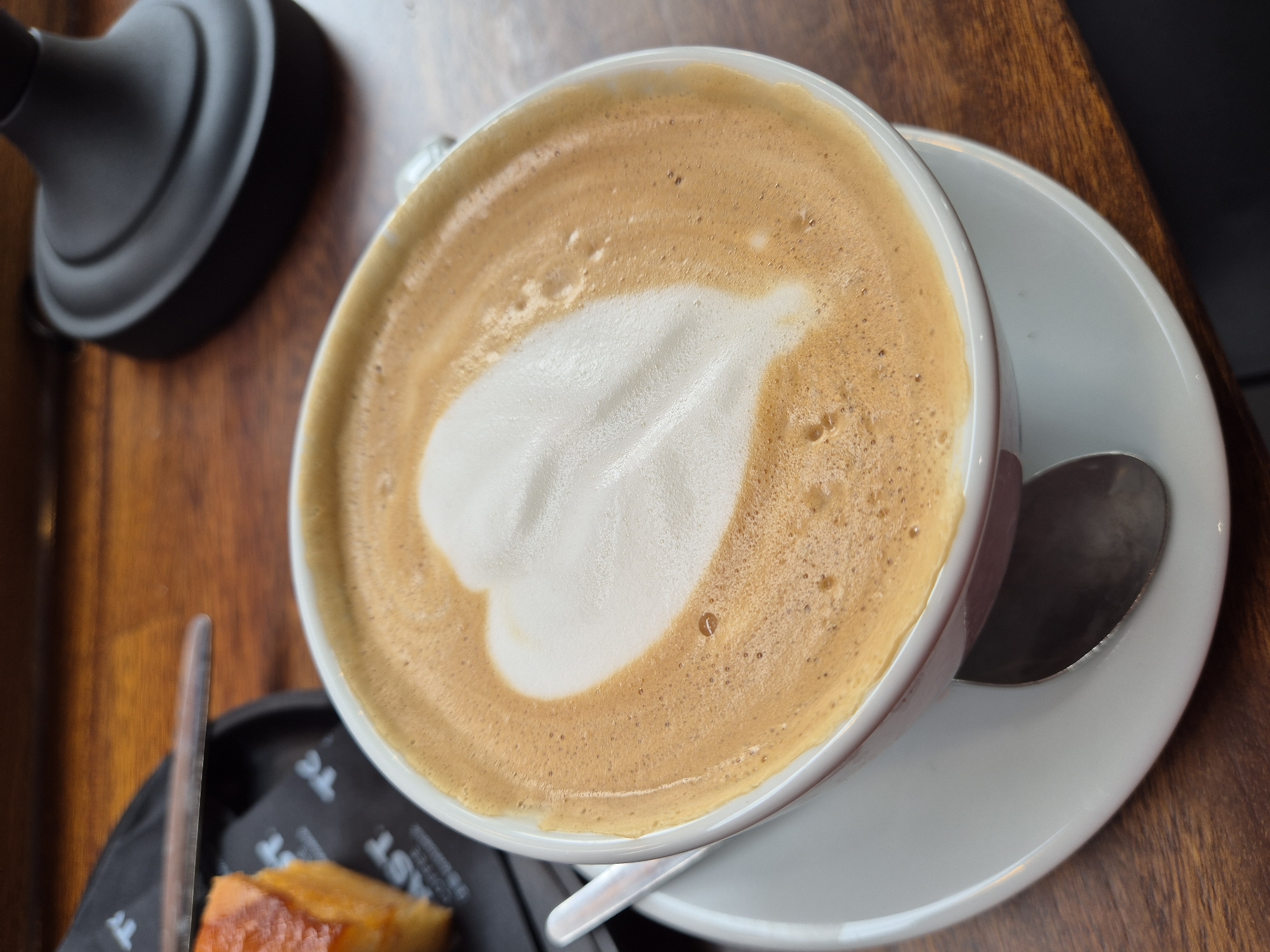 A close-up, high-angle shot depicts a cappuccino in a white cup, resting on a white saucer, with a spoon on the right side. The cappuccino features latte art in the form of a leaf or heart made of white foam, set against a tan-colored, foamy coffee background. Part of a black lamp base is visible on the left side of the frame. In the lower-left corner, a black rectangular plate holds what appears to be a pastry, partially visible, along with a piece of branded packaging. The background consists of a dark brown wooden surface. The image lacks specific details that definitively identify the location in West Suffolk, United Kingdom.