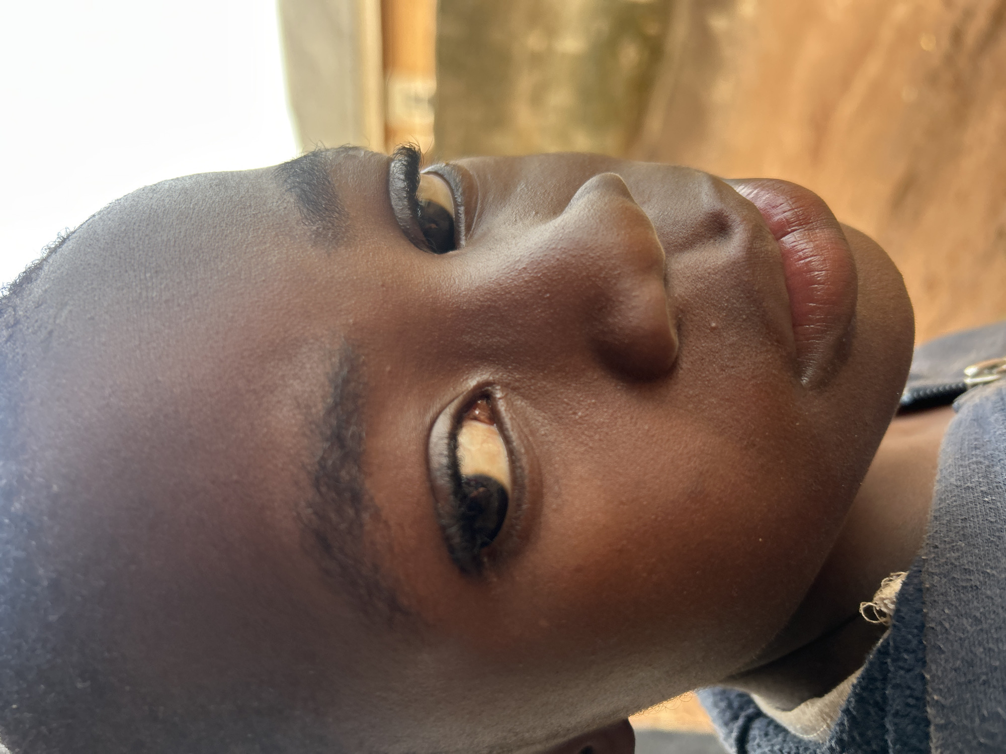 This close-up, low-angle portrait features a person with a dark skin tone, presented in a profile or three-quarter view, looking upwards. The frame primarily focuses on the individual's face, specifically the right side of their head, encompassing their eye, nose, and lips.
The person's eye is partially visible, with the pupil rolled upwards, exposing a significant portion of the sclera which appears yellowish-white. Long, dark eyelashes are prominent, framing the eye. A thick, dark eyebrow is also visible. The nose has a distinct, well-defined bridge and nostril. The lips are full and appear slightly parted, revealing a reddish-pink hue. The skin exhibits visible texture.
The individual is wearing a dark blue or grey hooded garment, visible around their neck and shoulder on the right side of the image. The background is softly blurred with bright, warm, and earthy tones, suggesting an indoor setting or a sheltered outdoor area with natural light. The lighting indicates it is daytime. There is no visible text in the image. The upward gaze gives the expression an ambiguous quality, potentially suggesting contemplation, weariness, or a specific artistic pose.