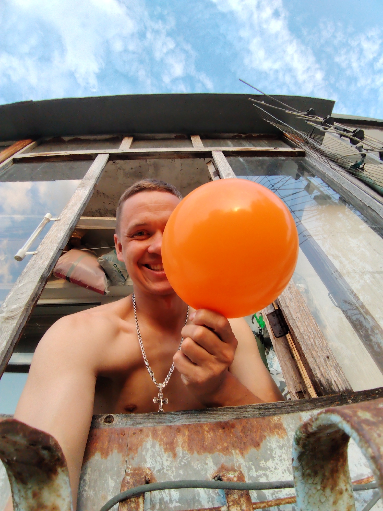 A shirtless man with a silver chain and cross is smiling and holding an orange balloon. He is standing in an open window with rusty metal framing. Outside the window, there is another window pane and a TV antenna. The sky is visible with some clouds. Based on the architectural features of the building and the presence of a specific type of TV antenna, this image is most likely taken in Chaikovsky, Russia.
