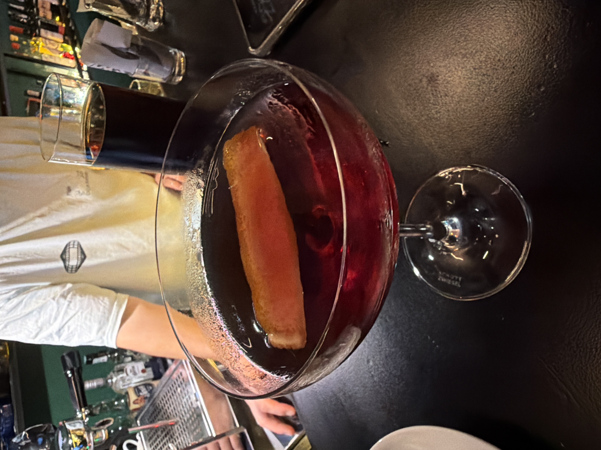 This image captures a close-up, overhead view of a bar scene in Budva, Montenegro, likely during the evening due to the dim lighting. The main subject is a large, clear cocktail glass holding a dark red beverage, possibly a cocktail, with a thick, rectangular orange-yellow garnish floating within it. To the left and slightly behind, another glass filled with a dark liquid, possibly a stout beer or dark soda, is visible. The base of the cocktail glass clearly shows the brand "SCHOTT ZWIESEL."
The drinks rest on a dark, textured surface, likely a bar counter. A person, possibly a bartender or server, is partially visible on the left side of the frame, wearing a light-colored shirt with a dark, stylized logo (resembling a diamond or shield shape) on it. Their arm is extended towards the drinks. Another person's hand is visible on the bar surface towards the bottom center.
In the background, typical bar elements are discernible: various bottles of spirits, metal bar equipment, and taps. A small glass containing a rolled-up napkin or paper is also visible. The setting conveys a casual, indoor environment, typical of a pub or restaurant.