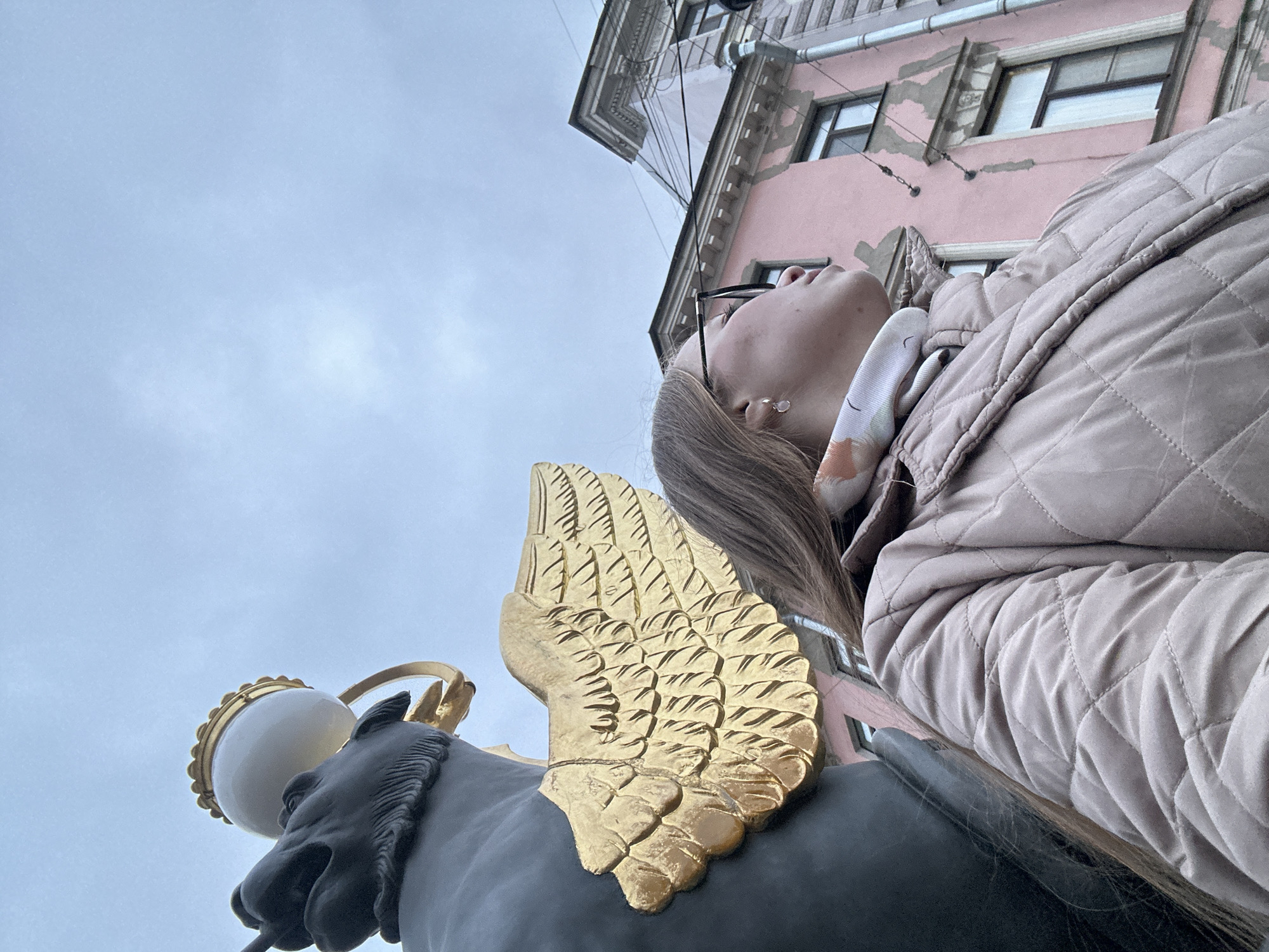 The image presents a low-angle perspective, looking upwards towards a sky, a building, and two primary subjects: a person and a decorative statue.
In the foreground and midground, a statue of a horse's head is visible, crafted from a dark material, possibly bronze or iron. It features a prominent, intricately detailed golden wing extending from its side, and an ornate, spherical white lamp with golden trimmings positioned above its head.
Adjacent to the statue, a person, likely a woman, is shown from the chin up in profile, looking upwards. She has light brown hair, is wearing glasses, and an earring is visible on her ear. Her attire includes a light beige or grey quilted jacket and a subtly patterned scarf. Her gaze is directed upwards, suggesting she is observing the statue, the building, or the sky.
The background features a distinct, pale pink building with multiple windows and classical architectural details such as cornices and moldings, indicative of historical European urban architecture. Thin wires are visible against the building facade and the sky. The sky itself is overcast and light blue-grey, suggesting daytime with diffuse lighting.
The overall scene captures a moment of observation amidst Saint Petersburg's urban landscape, with the ornate statue and distinctive building contributing to the city's rich architectural character. No visible text is present in the image.