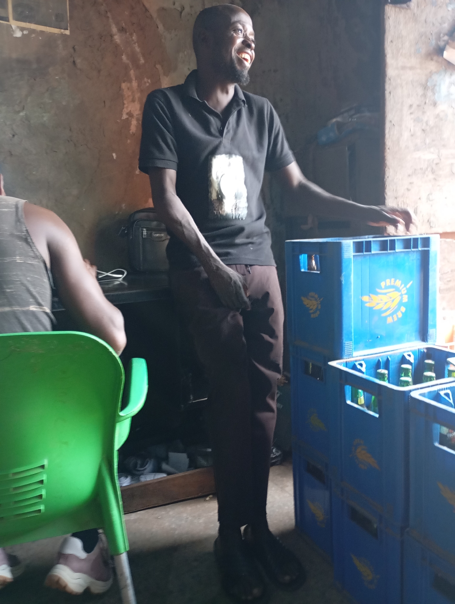 The image captures a man standing in what appears to be a small, rustic establishment, possibly a bar or shop, in Shendam, Nigeria. The man, who is the primary subject, is wearing a dark polo shirt and brown trousers. He has a beard and is smiling broadly, looking towards his right. His right hand is resting on a stack of blue plastic crates, one of which is labeled "PREMIUM BREW" with a yellow logo of a wheat stalk. Another crate, partially visible in the foreground, also contains green glass bottles, presumably filled with beverages.
To the man's left, another person's back is visible. This individual is wearing a grey tank top with horizontal stripes and is seated on a bright green plastic chair. The chair's design is functional, with horizontal slats on the back. Beneath the chair, two feet wearing pink and white sneakers are visible.
The background of the scene suggests an indoor environment with rough, textured walls, possibly made of mud or unplastered brick, typical of some rural or semi-urban structures in Nigeria. There is a black bag or satchel resting on a surface behind the standing man. The lighting in the image is dim, with natural light filtering in from the right, creating shadows and highlighting certain areas. The overall impression is of a casual, possibly local gathering place.