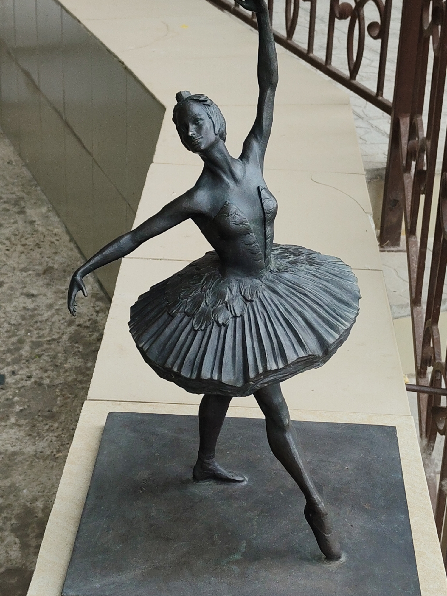 The image shows a dark bronze sculpture of a ballerina in a dynamic pose. The ballerina is wearing a tutu and is en pointe, with her arms outstretched. She has a focused expression on her face. The sculpture is mounted on a dark rectangular base, which rests on a light-colored tiled surface. In the background, there is a railing with decorative metalwork and a wall with a grid pattern. The lighting suggests it is daytime, with shadows cast by the sculpture and the railing. The overall scene is outdoor, likely in a public space or garden.