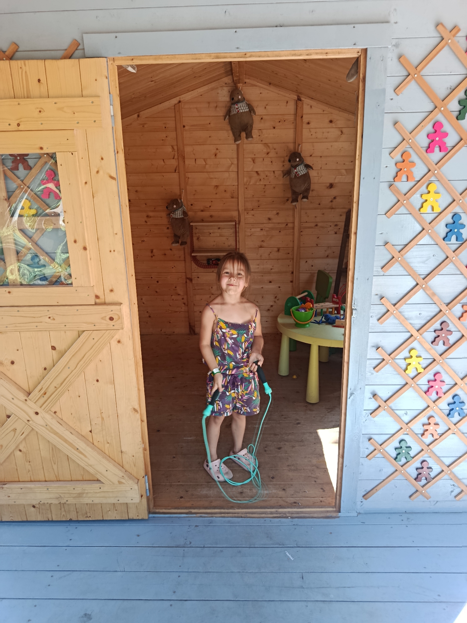 A young girl stands in the doorway of a wooden playhouse, holding a jump rope. She is wearing a floral print dress and pink shoes. The playhouse has wooden walls and a wooden floor. Inside the playhouse, there is a table with toys and some stuffed animals hanging from the walls. The exterior of the playhouse is decorated with a lattice pattern featuring colorful figures. The playhouse appears to be located outdoors in a yard, possibly in Upeslejas, Latvia, based on its design and the surrounding environment.