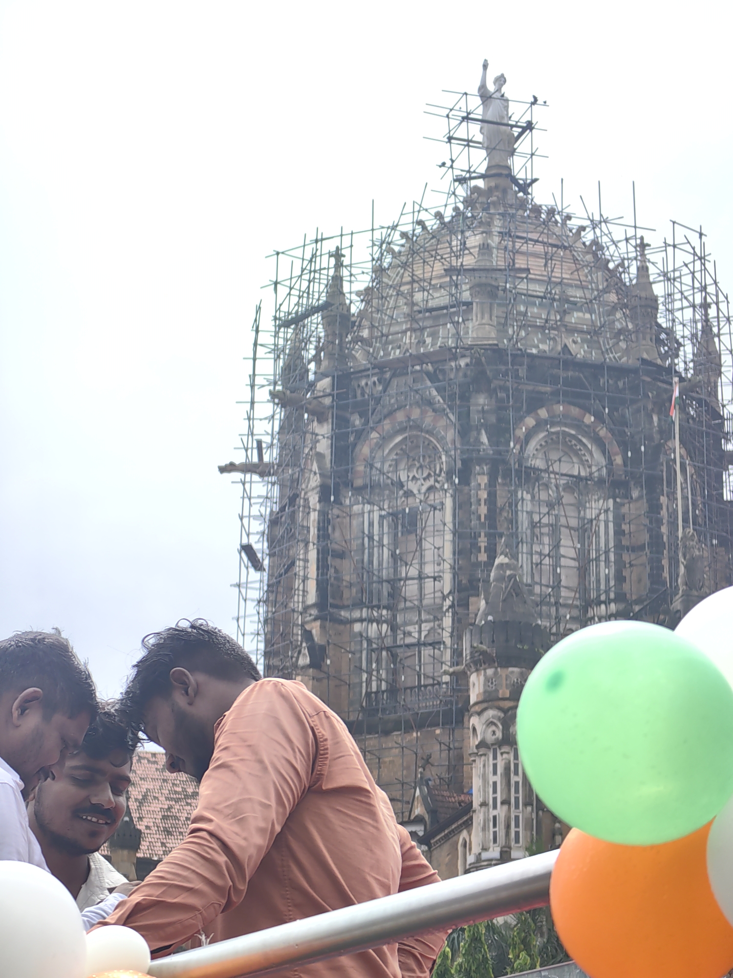 In Mumbai, India, Chhatrapati Shivaji Maharaj Terminus, a UNESCO World Heritage Site, is partially covered in scaffolding. In the foreground, a group of young men are visible, with three of them in focus. One man in an orange shirt is leaning forward, looking down at something out of frame, while two others stand beside him, one in a white shirt and the other in a light-colored shirt with a mustache. In the lower right corner, several balloons are present, including one green, one orange, and one white. The sky is overcast.