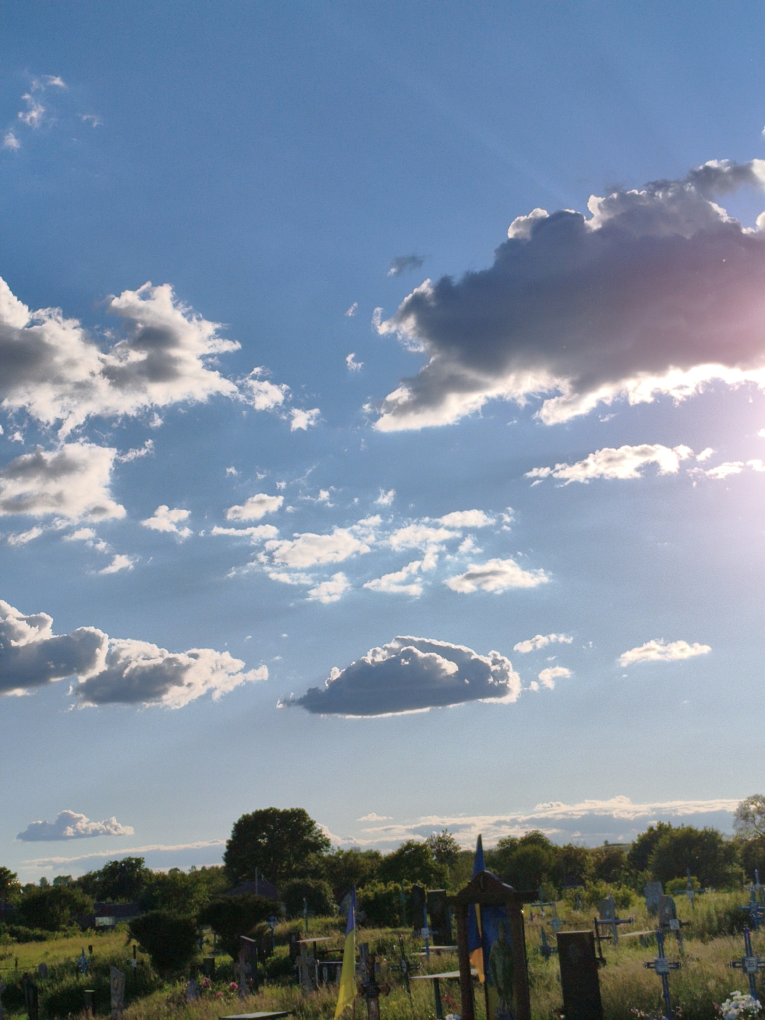 The image shows a cemetery under a blue sky with scattered clouds. The location is Unknown, Unknown. Gravestones and crosses are visible amidst overgrown grass and vegetation. In the background are trees and a few buildings. The sky is predominantly blue, with varying cloud formations ranging from fluffy white to darker, more ominous shapes. The sun is shining, creating a bright glare in the upper right of the image.