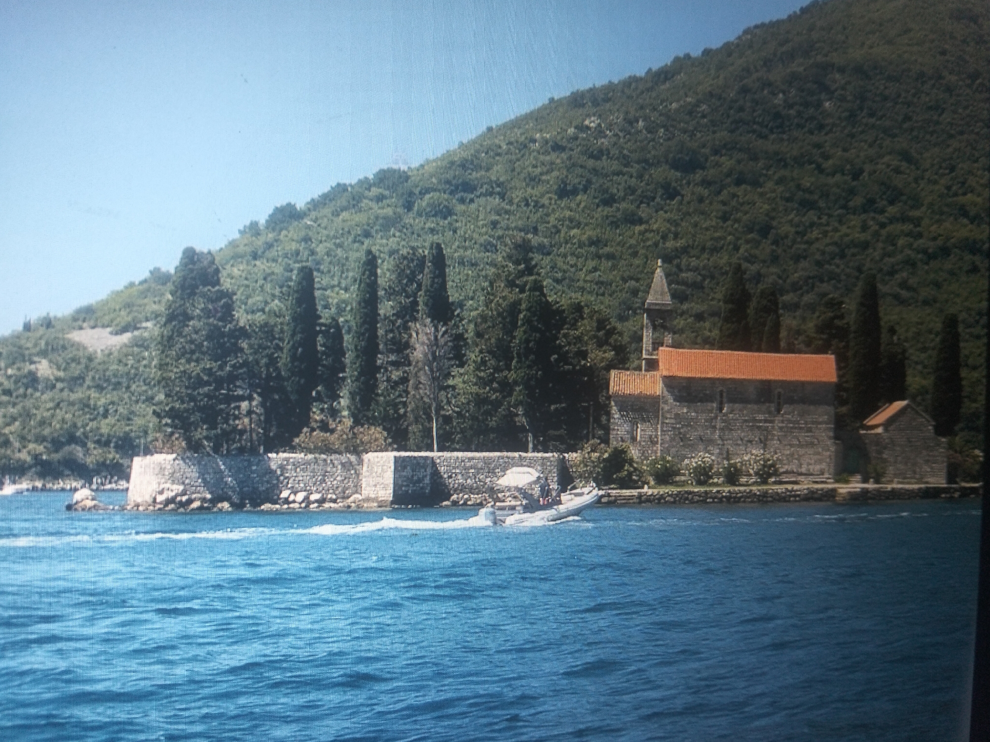 The image captures a scenic view of a coastal area with a lush green mountain forming the backdrop. In the foreground, a body of blue water reflects the clear sky. A white speedboat is seen moving across the water, creating a wake. Along the shoreline, a stone wall encloses a small island with a building complex. The complex appears to be a religious structure, possibly a monastery, with a prominent bell tower and a red-tiled roof. The surrounding vegetation is dense, with trees and bushes dotting the landscape. The overall scene depicts a tranquil and picturesque setting.