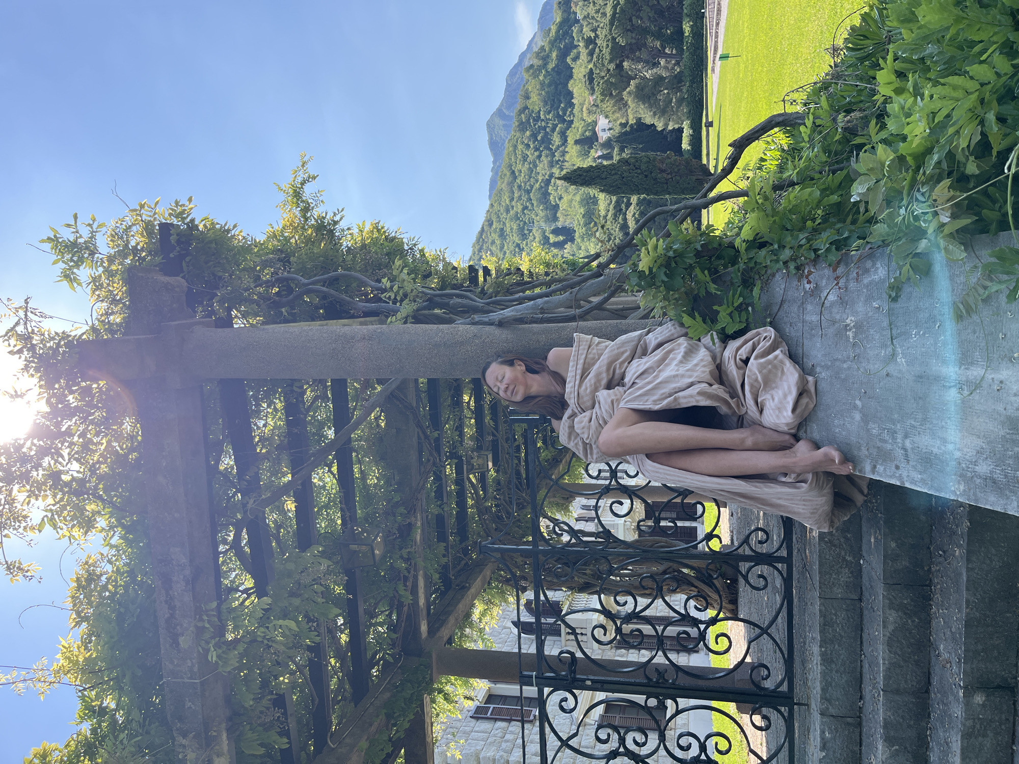 An outdoor, sunlit scene depicts a woman resting peacefully on a stone ledge, positioned horizontally within a picturesque garden setting. The woman, with dark hair, lies on her side, covered in a light-colored, possibly striped, fabric that drapes around her body, leaving her bare arms and lower legs visible. Her eyes are closed, suggesting sleep or deep relaxation.
She is situated on a light grey stone wall or ledge, beneath a rustic pergola or trellis structure made of thick wooden beams, which is heavily overgrown with lush green vines and foliage. Sunlight streams through the leaves, creating bright highlights and some lens flare on the left side of the image. Below the ledge and to the right, a decorative black wrought iron gate with intricate scrollwork is visible, alongside grey stone steps leading downwards. Behind the gate, a stone building with dark window shutters can be partially seen.
In the background, towering, tree-covered mountains rise under a clear, bright blue sky, indicating good weather. To the far right, a manicured green lawn stretches out. The overall atmosphere is serene and tranquil, enhanced by the natural surroundings and the woman's peaceful repose. The scene evokes a sense of calm and natural beauty, consistent with a cultivated garden in a mountainous region like Sveti Stefan, Montenegro. No visible text is present.