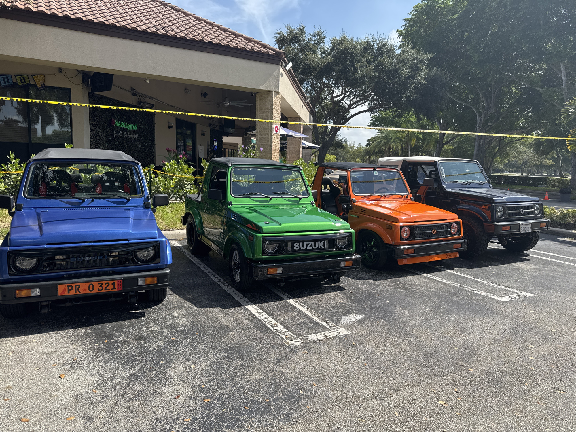Four Suzuki Samurai vehicles are parked in a lot in front of a building with a tiled roof in Coral Springs, Florida. The vehicles are blue, green, orange, and black. A yellow caution tape is strung across the front of the building. The building appears to be a restaurant, with a sign that says "MARGARITAS" visible. The sky is blue with some clouds.