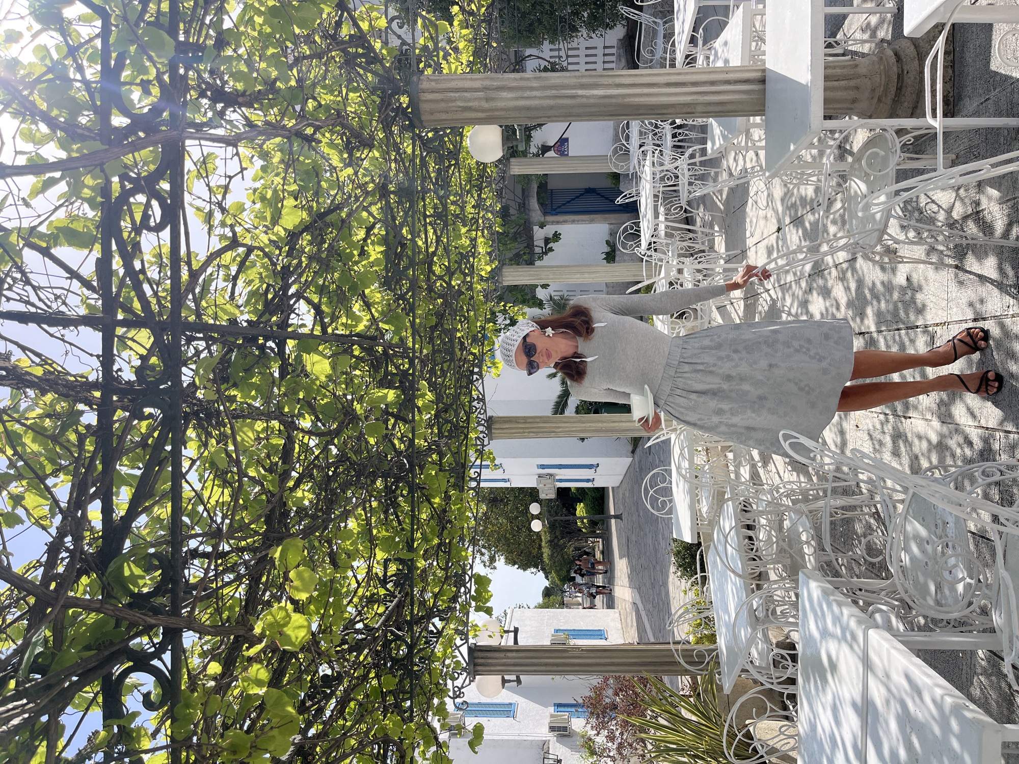 A woman stands on a sunlit outdoor patio, lined with white metal chairs and tables, framed by a leafy arbor supported by stone pillars. She wears a gray dress, a white head covering, and dark sunglasses. The background includes white buildings with blue shutters, typical of Mediterranean architecture. In the distance, other people can be seen walking along a paved pathway. The location is likely in or around the Old Town of Budva, Montenegro, characterized by its historic buildings and outdoor cafes.