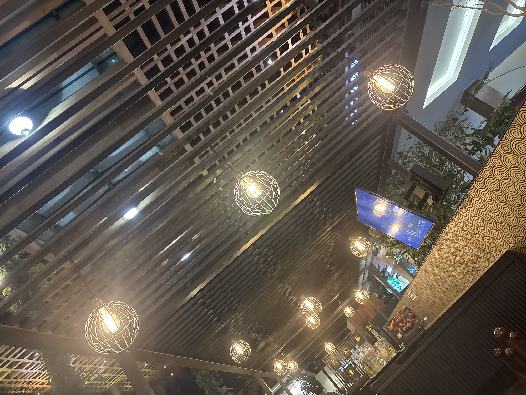 The image captures the interior of a PF Chang's restaurant, likely situated within a modern commercial complex such as Angelópolis Lifestyle Center in Puebla, Mexico. The perspective is steeply angled upwards, showcasing a ceiling constructed from a dense array of dark, horizontal slats, possibly wood or metal. Numerous spherical pendant lights, each featuring a wire cage design encasing an incandescent-style filament bulb, hang at various heights from this slatted structure, casting a warm, yellowish glow across the scene.
Beyond the primary slat grid, additional architectural elements are visible, including areas with a finer lattice pattern and reflections of bright, possibly recessed, overhead lighting. In the background, geometric forms of white and blue construction are apparent, suggesting an expansive or multi-level interior. Green foliage from potted plants is interspersed throughout the background, adding organic elements to the commercial setting.
In the lower left quadrant, a dark-colored counter with a decorative surface patterned with concentric circles in brown and black is prominent. Resting on this counter are several glass bottles containing liquids. A small, black menu holder on the counter clearly displays the text "PF CHANG'S" at the top and "PEKING DUCK" below. Behind the counter, multiple rectangular signs adorned with Chinese characters are mounted. The characters "大家" (Dàjiā - everyone/all), "喜" (Xǐ - joy/happiness), and "精" (Jīng - essence/spirit) are discernible.
To the right, a large, vertically oriented screen displays abstract blue and white graphics, which appear to be reflections of the ambient lighting and cage pendants. Through a transparent barrier or reflection in the background, a green circular logo, consistent with a Starbucks brand mark, is visible. In the extreme lower left corner, a human hand, possibly male, adorned with rings, rests on the dark, ridged edge of the counter.