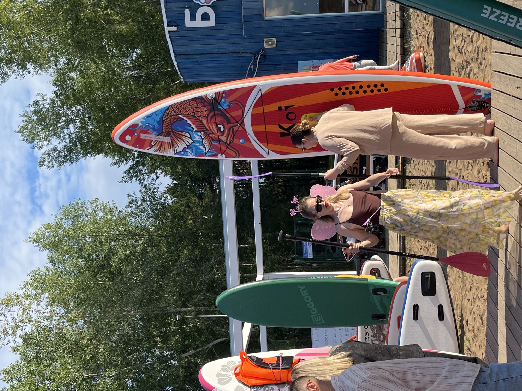 The image shows a group of people gathered around a collection of stand-up paddleboards (SUPs) on a sandy area. In the foreground, a woman with pink fairy wings and a crown is holding a paddle, and another woman in a beige outfit is standing behind a colorful SUP decorated with a koi fish design. Several other SUPs are visible, including a green one with the text "EGGORY" and a white and blue one. In the background, there is a blue building with the number "5" visible on it, and lush green trees under a bright blue sky with white clouds. The scene appears to be a sunny day, likely in the late morning or afternoon, given the lighting. The overall atmosphere seems relaxed and perhaps celebratory, with the fairy costume suggesting a fun or themed event. Visible text includes "EGGORY," "Koi," and "REEZE," along with Russian text that translates to "Chaikovsky Krai."
