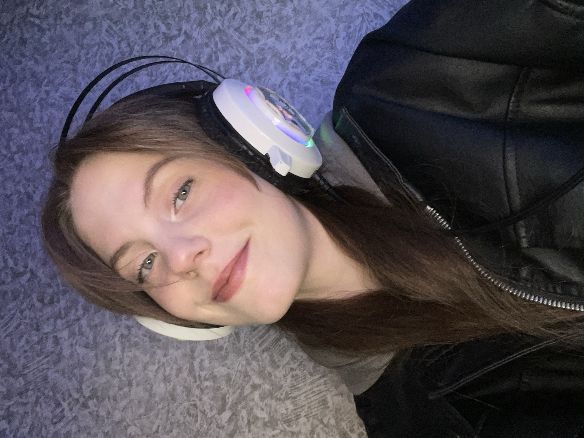 A young woman is seen in a close-up, angled selfie. She is wearing headphones with white earcups and colorful lights and a black leather jacket over a gray t-shirt. The background appears to be a patterned carpet or rug. The lighting is somewhat dim and possibly artificial. There is no indication of the city of Фоки, Russia in this image.