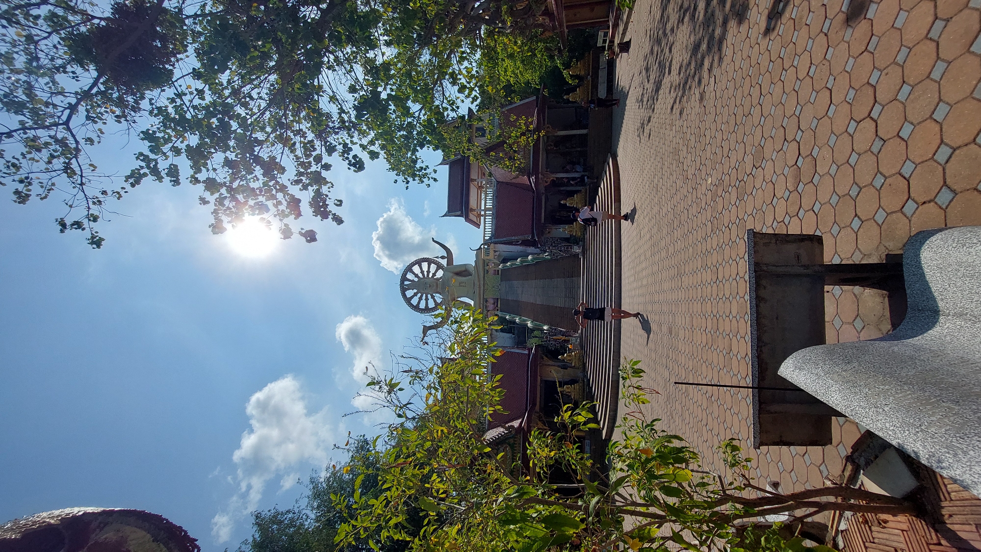 The image shows a sunny outdoor scene in Baan Plai Laem, Thailand, featuring a temple complex. In the center is a large Buddha statue with a Dharmachakra wheel. Two people walk up stairs that lead to the statue. The ground is covered with a patterned brick walkway. Vegetation is visible, including trees with green foliage, partially obscuring the sky, which is blue with scattered clouds. The sun is positioned high in the sky. A stone bench is in the foreground.