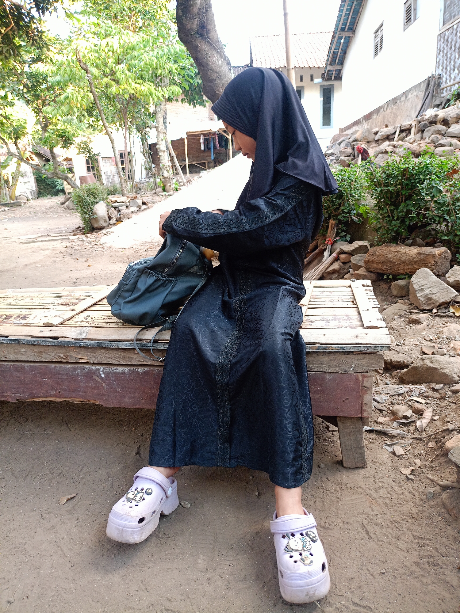In a rural outdoor setting in Jelbuk, Indonesia, a young person wearing a black hijab and a patterned black abaya is seated on a weathered wooden bench. Their feet, clad in light purple platform clogs decorated with charms, rest on the dirt ground. The individual is focused on a dark green backpack, which they are handling. The background reveals a rustic environment with a dirt path, scattered rocks, greenery, trees, and simple buildings constructed with wood and white walls.