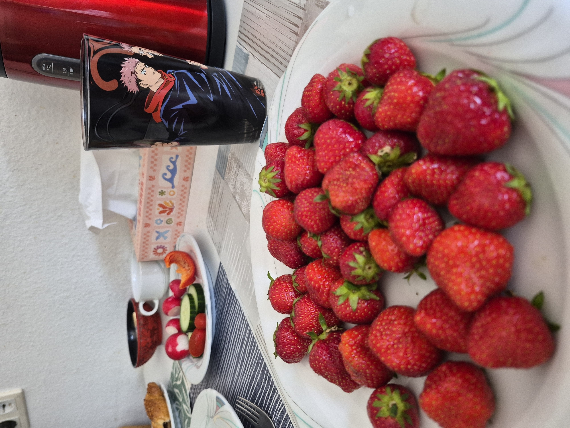 A close-up, high-angle shot shows a table with a variety of food items, including a plate of strawberries, a plate of vegetables, and a red mug with an anime character printed on it. The strawberries are plump and bright red, filling a white bowl. To the left of the strawberries, a small white plate holds sliced radishes, cucumber, and a piece of red bell pepper. A red mug with a black base and an anime character is positioned behind the plates of food. The mug features a character with pink hair and a blue jacket. A red appliance, possibly a kettle or toaster, is visible in the background. The table appears to be set for a meal or snack, with a patterned placemat under the plates. The scene is brightly lit, suggesting it is daytime. No specific location cues are visible, and the overall mood is casual and domestic. The mug has some text printed on the side with indicators for volume settings.