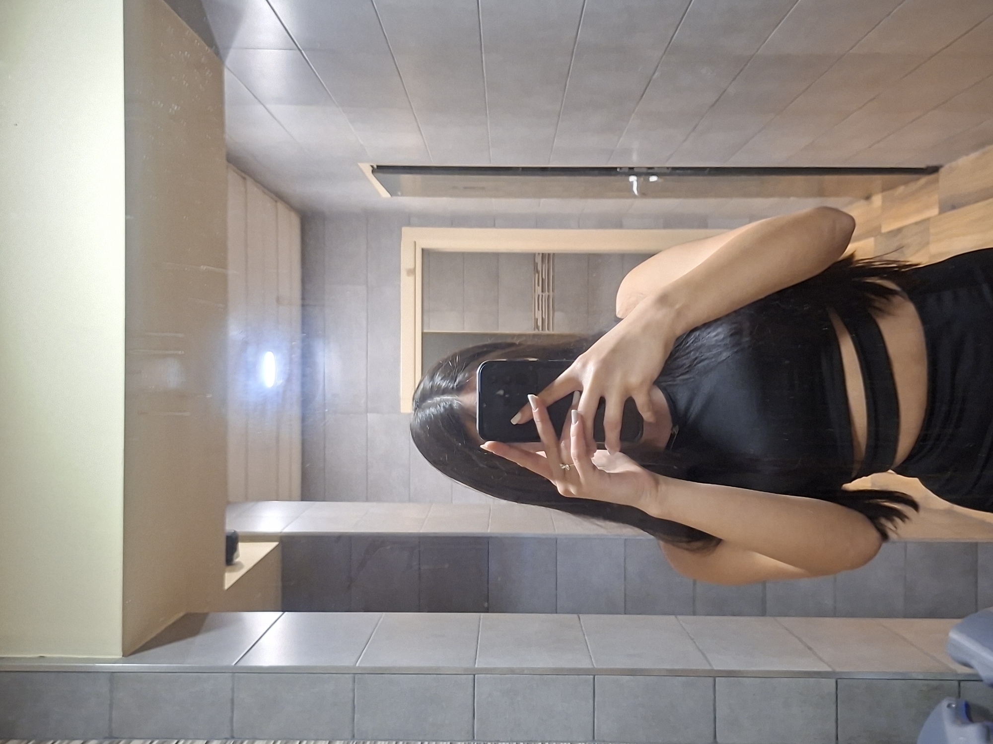 The image shows a woman taking a selfie in a bathroom. The woman has long, dark hair and is wearing a black outfit with cutouts in the back. She is holding a smartphone in her hands to take the picture. The bathroom has gray tiled walls and floors, and there is a mirror reflecting the scene. A beige-colored structure is visible in the background, potentially part of the bathroom fixtures or cabinetry. The exact location in the city of Pleven, Bulgaria, is indeterminable from the given image.