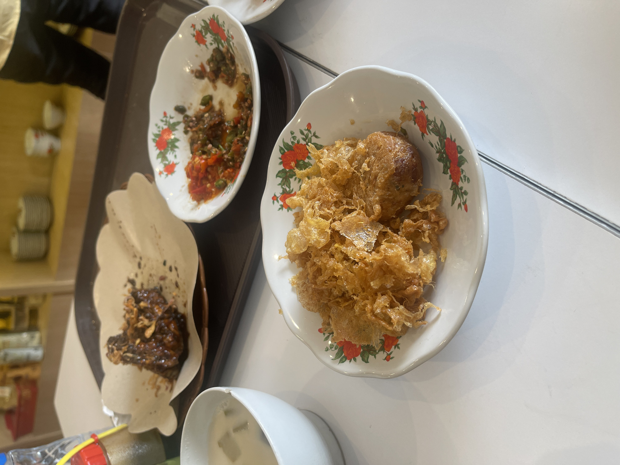 An overhead shot shows a white table laden with Indonesian food. On the left, a white bowl contains a light-colored soup with green garnish. In the center, a plate holds fried, crispy food that appears to be egg or a similar batter. To the right, another small white bowl contains a spicy red chili and vegetable condiment. Further back, a larger plate is adorned with a dark glazed meat dish, possibly chicken or beef, topped with fried shallots, served on a piece of textured brown paper. The food is arranged on a dark brown tray, suggesting it was served from a restaurant. The background shows shelves with stacked plates and bottles, indicating a dining establishment in Medan, Indonesia.