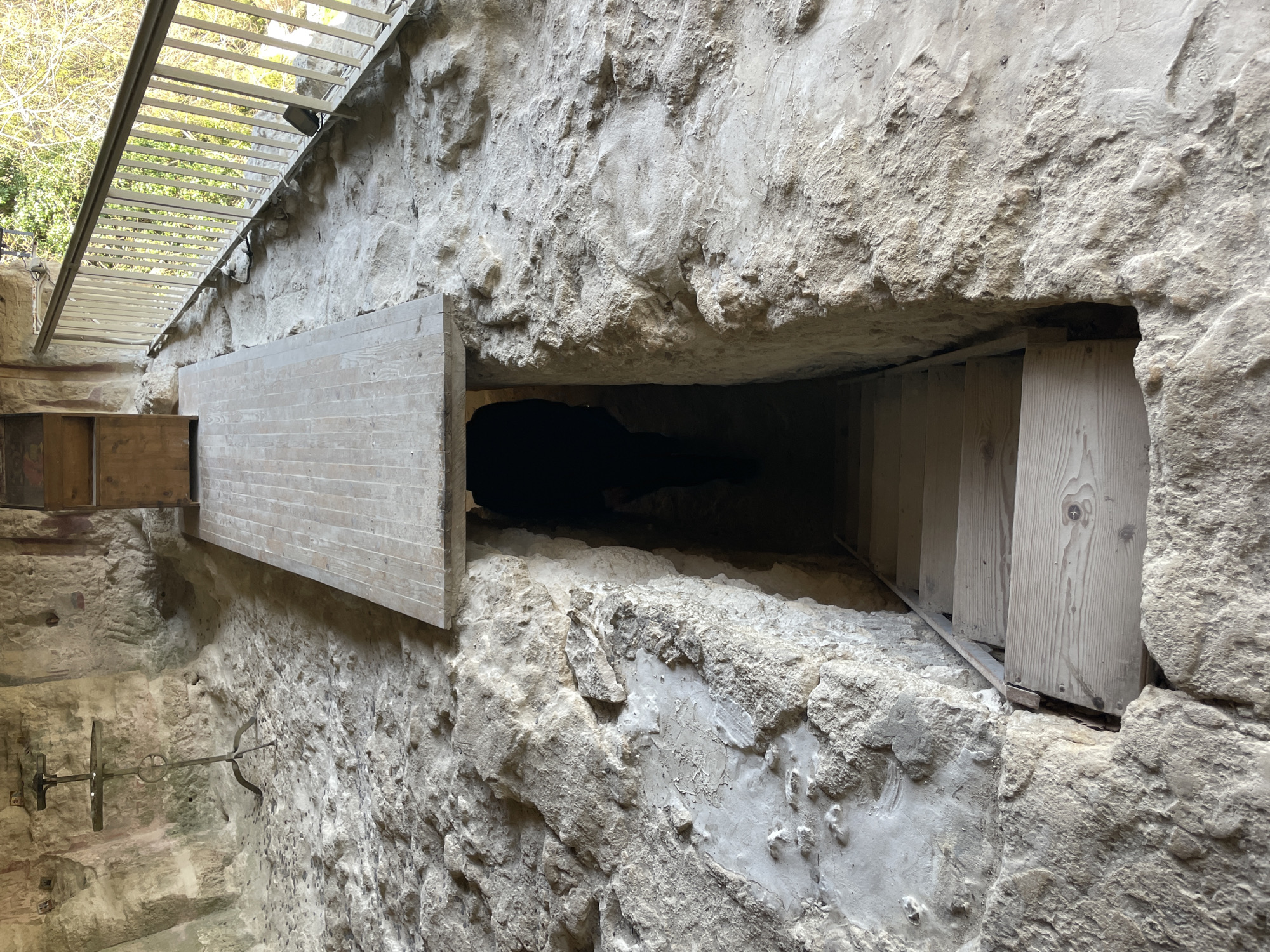 The image depicts a rough, possibly ancient stone wall with a narrow, dark opening. The opening is partially covered by wooden planks on both sides, with one section appearing more modern. A metal framework structure is visible above the wall. The scene might be located in Varna, Bulgaria, based on the prompt's instructions.