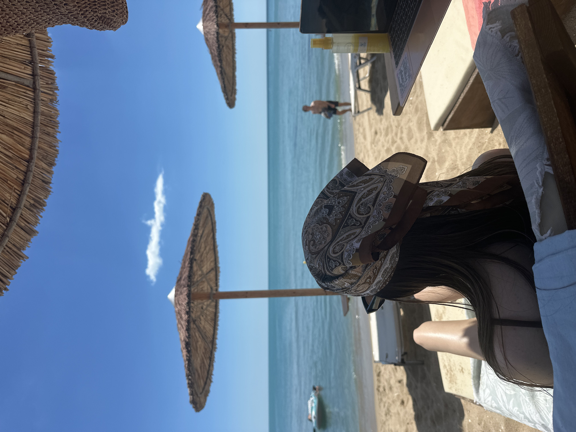 This image depicts a sun-drenched beach scene, likely on one of Varna, Bulgaria's numerous beaches, such as those near the Sea Garden. The perspective is from behind a person relaxing on a sun lounger, facing the Black Sea.
The foreground features the back of a person wearing a patterned headscarf and sunglasses. Their legs are extended on the lounger, which is covered with white and patterned towels. To the right, a laptop and a yellow spray bottle are visible on a wooden table, suggesting work or leisure activities.
In the middle ground, the sandy beach is dotted with empty sun loungers and thatched umbrellas providing shade. A person, a male, stands near the water's edge, holding a bag. The calm, turquoise waters of the Black Sea stretch towards the horizon, meeting a clear blue sky with a few wispy clouds. A small inflatable object is visible in the water to the left. The overall atmosphere is one of relaxation and summer enjoyment.