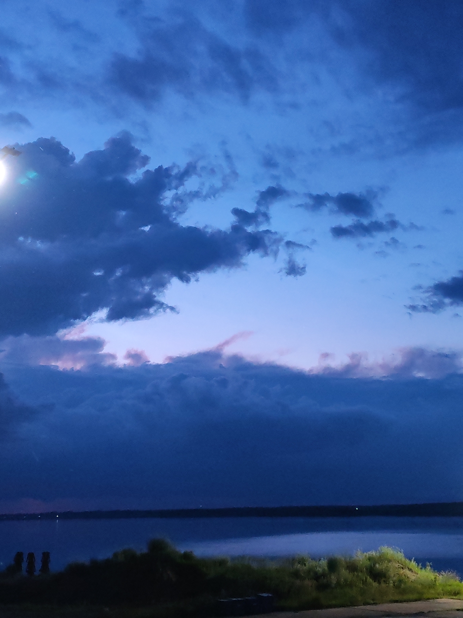 This is a vertical, medium-shot photograph taken at dusk, likely from a high vantage point overlooking a body of water. The sky dominates the upper two-thirds of the frame, filled with dark, heavy clouds tinged with shades of deep blue and purple. Towards the horizon, there is a faint band of lighter blue and pale pink, suggesting the last vestiges of daylight. A bright, almost blinding light source, likely a street lamp, is visible in the upper left corner, creating a lens flare and casting a soft glow on the surrounding clouds.
The lower third of the image depicts a dark, still body of water, possibly a river or lake, stretching across the width of the frame. A distant shoreline is visible as a dark, indistinct line. In the foreground, a strip of land is covered in dark, shadowy vegetation, appearing as a silhouetted mound. Three dark, vertical posts or pillars are faintly visible on the left side of the foreground, emerging from the vegetation. The overall mood is somber and tranquil, evoking a sense of twilight stillness. The location context of Rybinsk, Russia, suggests a potentially vast expanse of water typical of this region. There is no visible text in the image.