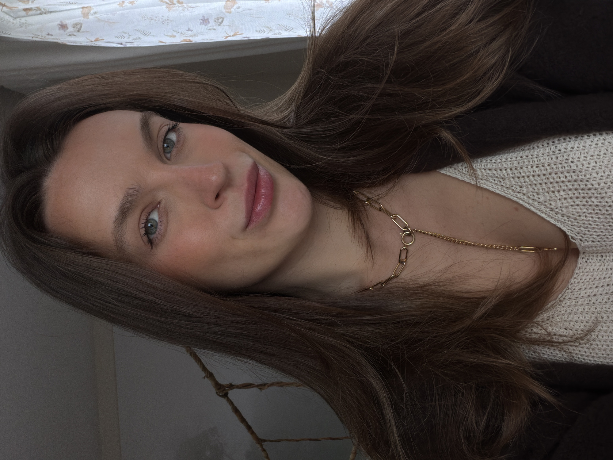 A close-up portrait features a woman with long, brunette hair, who is positioned at an angle. She is wearing a light-colored, textured sweater under a darker jacket or cardigan, and a gold chain necklace. The woman has fair skin, lightly blushed cheeks, green eyes, and pink lips. The background behind her is blurred, but a patterned surface, possibly a curtain or fabric, is visible in the upper left corner. A rope is visible in the lower left corner. The location in the city of Budva, Montenegro is not provided in the image.
