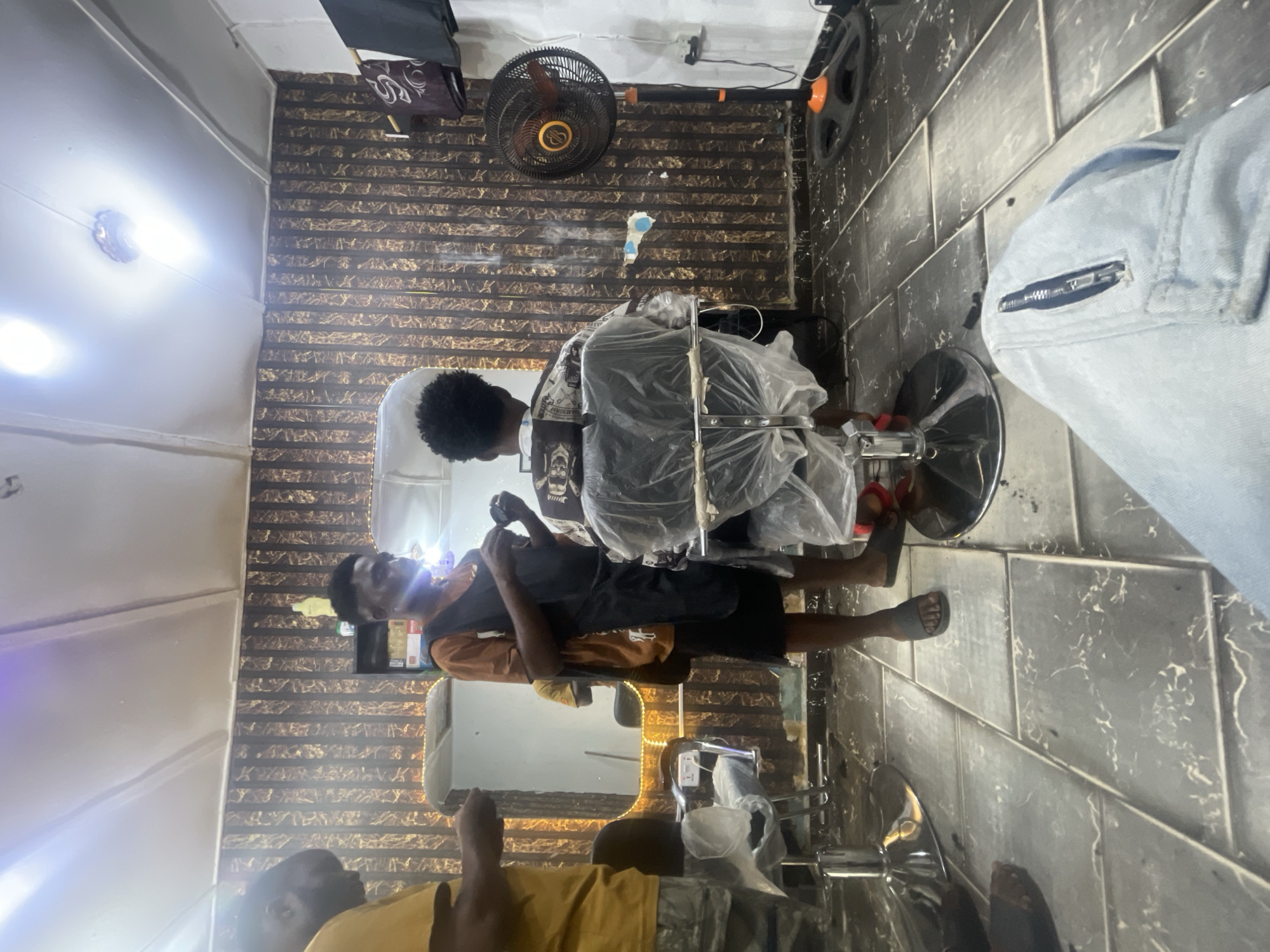The image captures a scene inside what appears to be a small barbershop, likely in Lokoja, Nigeria. The shop has a textured wall with horizontal lines, and a ceiling fan is mounted on the wall. Two men are visible; one is seated in a barber's chair covered in plastic, presumably to protect it from hair clippings. The second man, likely the barber, stands over the seated man, holding what appears to be cutting tools, as if in the middle of a haircut. Another individual wearing a yellow shirt is seen in the lower part of the image, potentially waiting for service. The floor is tiled with dark-colored tiles. The shop seems cramped, containing a mirror and barbering equipment.