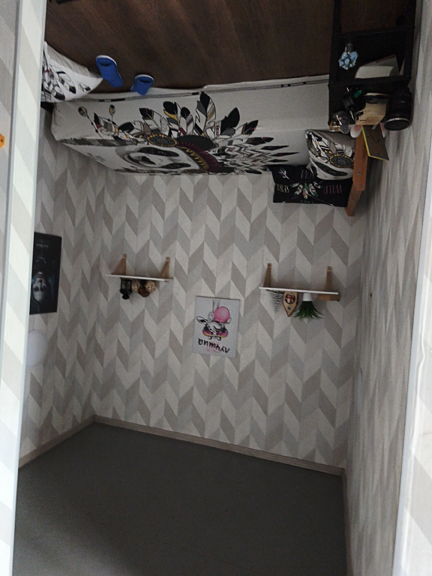 This media file captures an interior view of a bedroom or personal space, presented as if the image has been rotated 90 degrees clockwise.
**Subjects:** No people are visible in the image.
**Objects:** The main furniture piece is a bed, adorned with bedding featuring a distinctive dreamcatcher and feather design in black, white, and muted colors. A pillow with a similar design is visible, along with another black pillow that partially reads "WILD" (likely "WILD SPIRIT"). A pair of bright blue slippers rests on the bed. Adjacent to the bed is a dark bedside table or shelf unit, holding various small items including what appears to be a small green plant, a container, and other decorative objects.
The walls are covered in a geometric wallpaper with a chevron or herringbone pattern in shades of grey and white. Three framed pictures or posters are affixed to the wall: one on the left featuring a dark image, possibly a face, with faint text; a central poster depicting a pink cartoon character (possibly an elephant) with text below it that is difficult to fully decipher; and a third, partially obscured poster on the right, which seems to echo the dreamcatcher motif of the bedding. Two small, light-colored wooden floating shelves are mounted on the wall, each holding decorative items such as woven balls and another small green plant. The floor is covered with a dark grey carpet or rug, and a light-colored baseboard runs along the bottom of the walls. Parts of a doorframe or wall edge with vertical grey and white stripes are visible on the left and right, framing the scene.
**Scene/Setting:** The image depicts a corner of a bedroom, showcasing personal decor choices. The overall aesthetic is modern with touches of bohemian influence, particularly through the bedding design.
**Activity or event taking place:** No specific activity is occurring. The room appears tidy and arranged for daily living.
**Notable details:** The room's decor suggests a youthful or personalized space. The image's rotated orientation is a significant detail, making the bed appear vertical. The location context provided is Sevastopol, Ukraine, though no specific visual cues within the room directly confirm this.
**Visible text:** On the black pillow, "WILD" is clearly visible, likely part of "WILD SPIRIT." Text is also present on the central poster below the cartoon character, but it is not clearly legible.