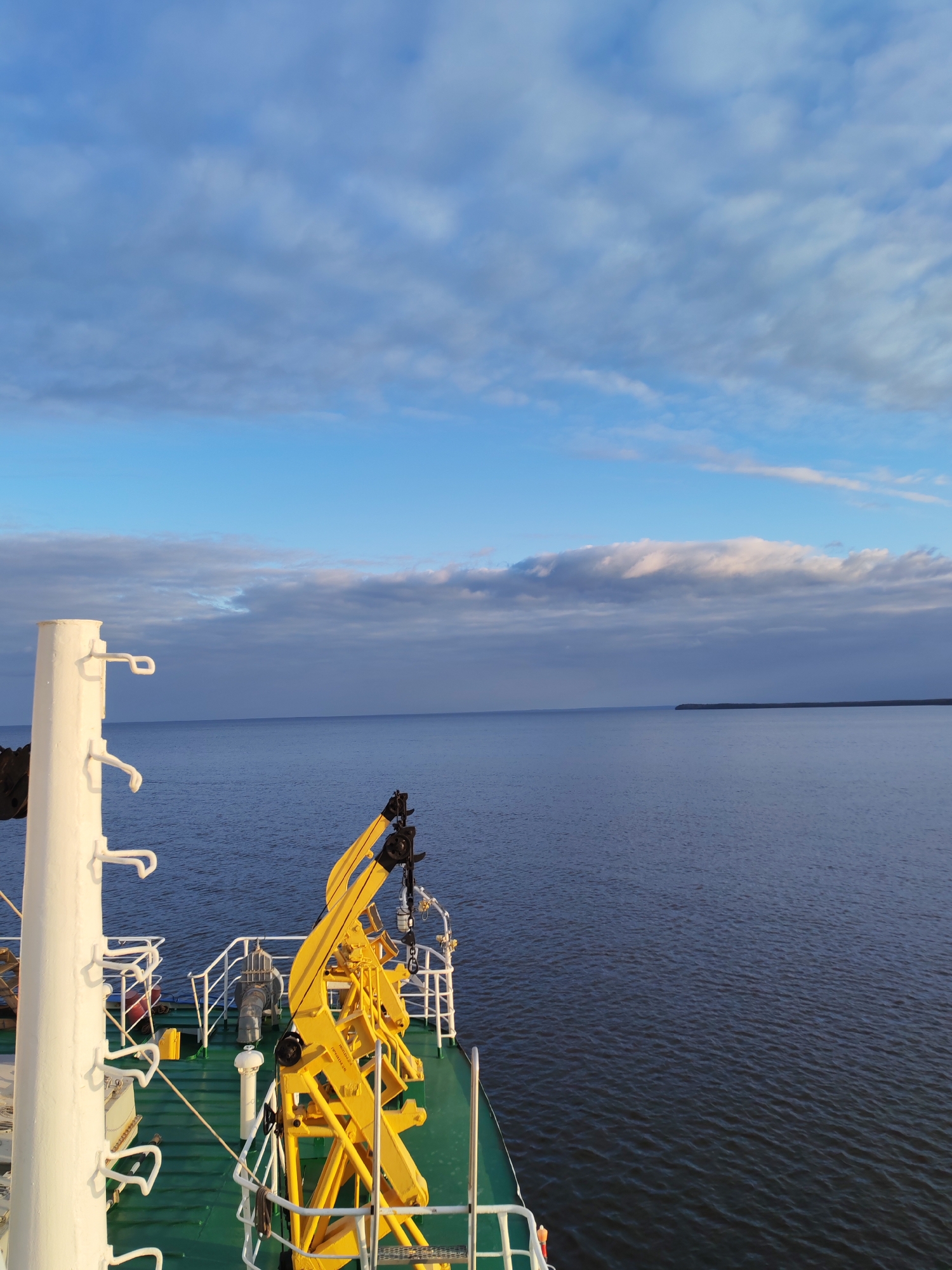 The image presents a serene maritime view from the deck of a ship, looking out over a calm body of water towards a distant landmass under a partly cloudy sky, likely in the vicinity of Shcheleiki, Russia.
In the foreground, part of the ship's green deck is visible. A prominent white mast or pole with multiple hook-like attachments stands on the left. Towards the center-right, a large, bright yellow crane structure, equipped with chains and hooks, dominates the deck machinery, implying industrial or cargo handling capabilities. White railings border the deck, curving gently along its edge. Beyond the crane, other pieces of equipment are discernible on the green deck, including what appears to be a winch or capstan.
The middle ground features a wide expanse of dark blue water, calm with slight ripples, stretching out to the horizon. A low, dark strip of land is visible in the far distance on the right, suggesting a coastline or island. The sky occupies the upper half of the frame, showcasing a mix of bright blue patches and various cloud formations, ranging from thin, wispy streaks to thicker, greyish masses.
The lighting indicates late morning or early afternoon, with clear, gentle sunlight. The overall atmosphere is one of tranquility and open space. No people are visible, and no discernible text can be read on any of the objects, although faint markings are present on the yellow crane. The scene depicts a ship either at sea or traversing a large body of water.