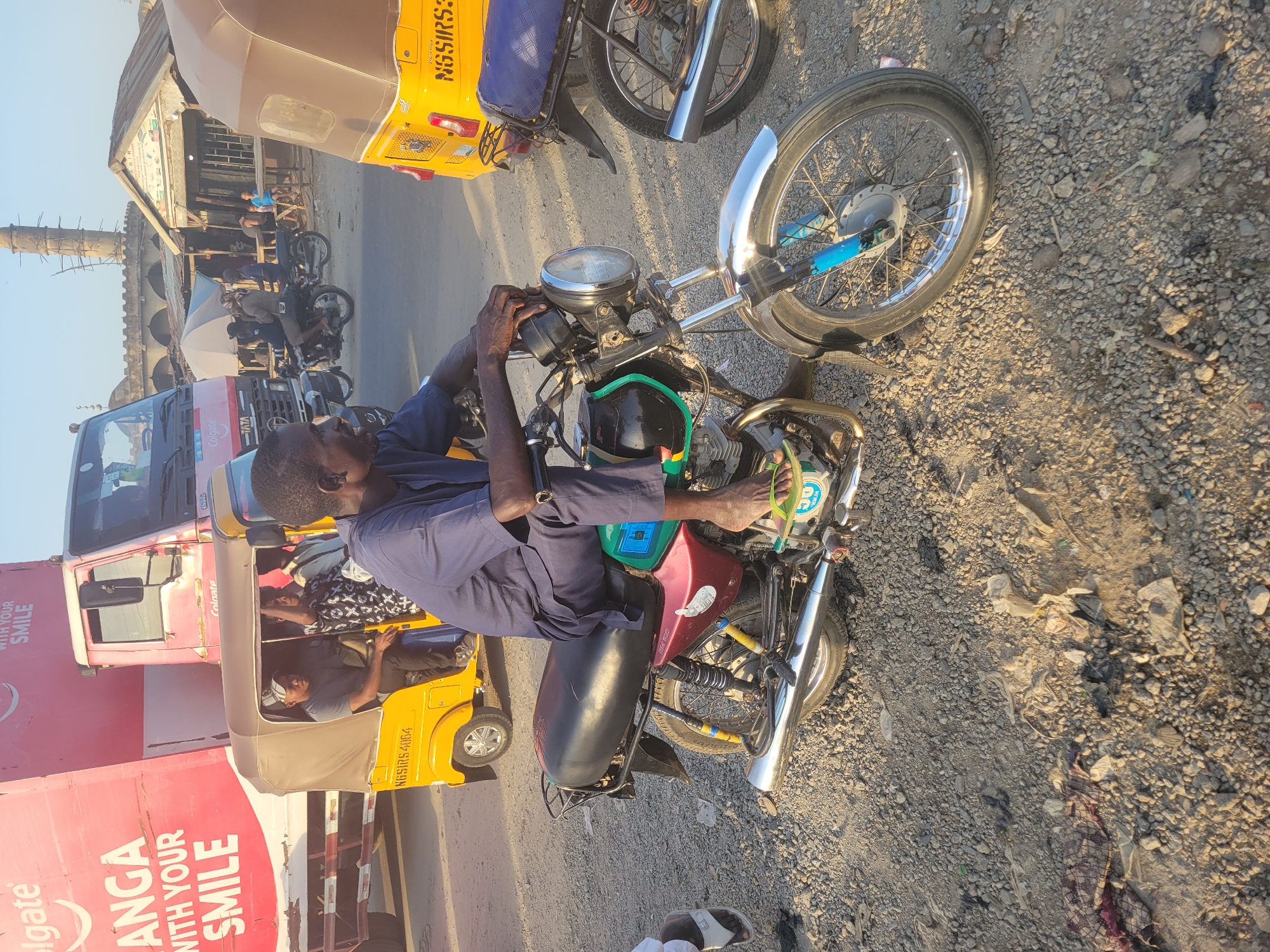 This media file captures a vibrant street scene in Madalla, Nigeria, during what appears to be late morning or early afternoon, characterized by bright sunlight and clear skies.
The primary subject is a man, dressed in a blue uniform, sitting on a red motorcycle and seemingly engaged in working on or inspecting it. His hands are near the handlebars and engine area. He is barefoot, with one foot visible resting on the motorcycle.
Around him, the scene is bustling with various forms of transportation and activity. To the man's left, a yellow rickshaw (tuk-tuk) with a beige canopy is parked or moving slowly, carrying at least two passengers. Another yellow rickshaw is partially visible further back. A large red truck, branded with "Colgate" and the slogan "ANGA WITH YOUR SMILE" (likely a local variation or language), dominates the left side of the frame, suggesting commercial activity. Several other motorcycles are visible in the mid-ground.
The background features a dusty, unpaved road and buildings. A prominent, tall, slender structure resembling a minaret indicates the presence of a mosque. A building with a corrugated metal roof is also seen, with some people sitting under a small umbrella nearby, suggesting a market or resting area. The ground is a mix of dirt and gravel, with some scattered litter.
Visible text includes "NASSIRS 4064" on the yellow rickshaw in the foreground, and "Colgate" and "ANGA WITH YOUR SMILE" on the red truck. The overall setting portrays a typical, active urban environment in a developing country, consistent with the provided location context of Madalla, Nigeria.