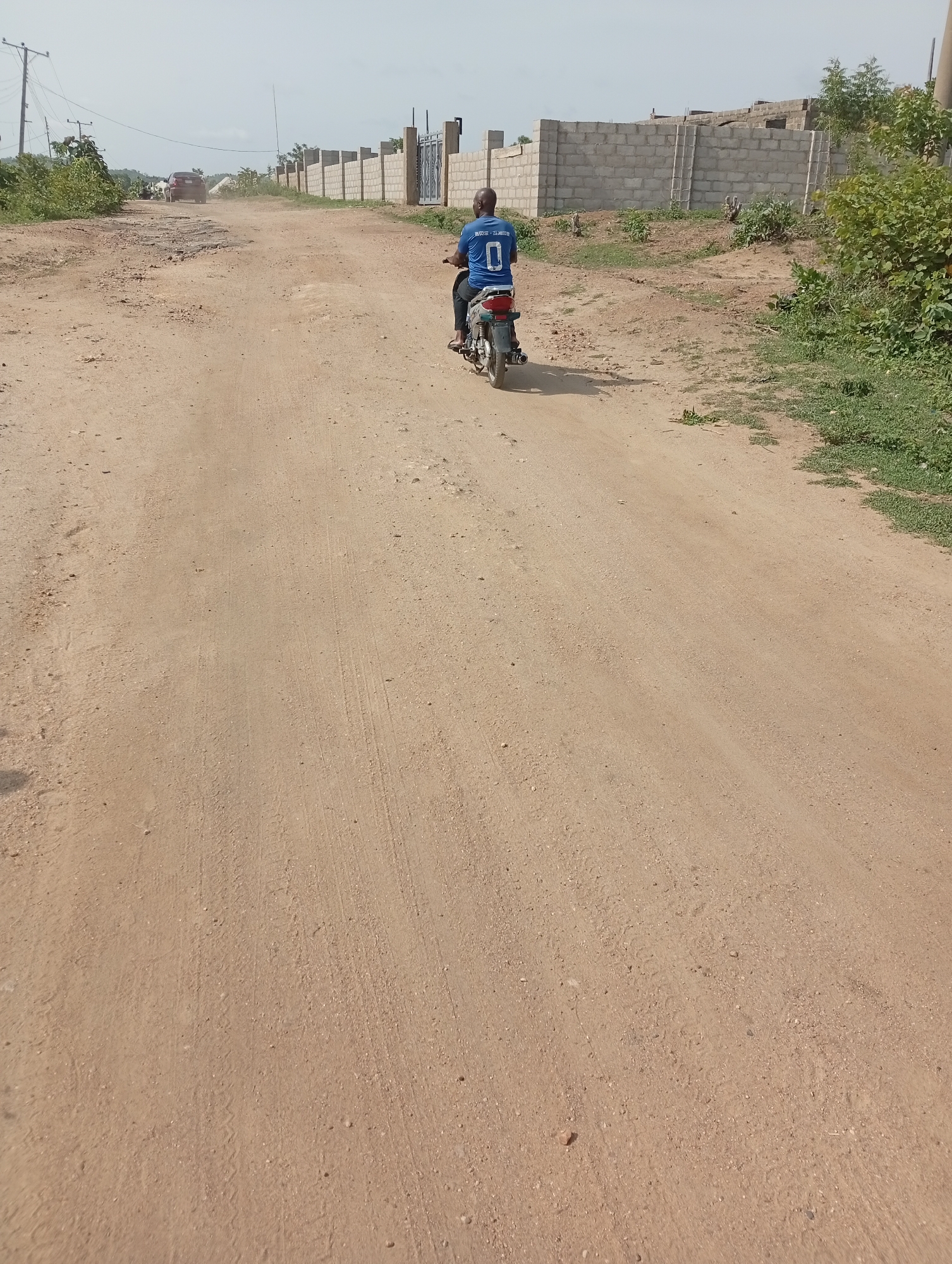 The image shows a man riding a motorcycle on a sandy road in Adankolo, Nigeria. He is wearing a blue shirt with the number "0" on the back. The road appears unpaved and dusty. A concrete block wall lines one side of the road, while vegetation grows on the other side. In the distance, a vehicle is kicking up dust. Overhead power lines are also visible in the distance.