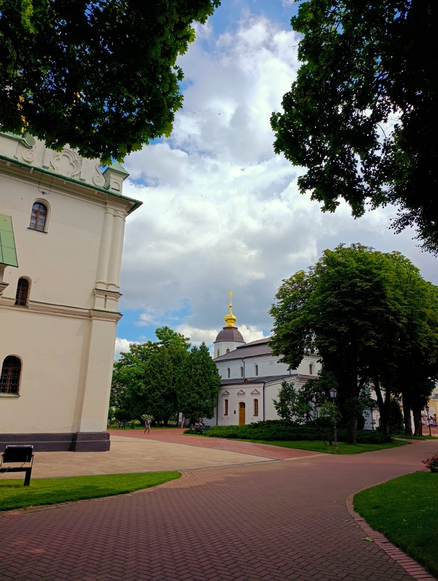 This is a medium-shot, vertical, outdoor photograph taken on a cloudy day in Kyiv, Ukraine. The scene depicts a courtyard area with pathways, green spaces, and prominent architectural structures.
On the left side of the frame, a large, pale yellow building with a green roof is partially visible. It features arched windows with dark wooden frames and decorative architectural elements near the roofline. To the right of this building, a smaller, white structure with a brown roof and a golden cross atop its dome is visible. This building also has arched windows and a yellow door. The architectural style suggests it is likely a church or part of a religious complex.
The foreground is dominated by a brick pathway with a herringbone pattern, which curves into the scene. Adjacent to the pathway are well-maintained green lawns.
A person, appearing as a distant figure, is walking on another pathway further into the scene. They are dressed in dark clothing. Several benches are visible in the background, some occupied by individuals.
Large, lush green trees frame the top of the image and flank the buildings, providing a natural contrast to the man-made structures. The sky is filled with dramatic, white and grey clouds, suggesting either late afternoon or an overcast day. The overall impression is one of a peaceful and historic setting. No visible text is present in the image.