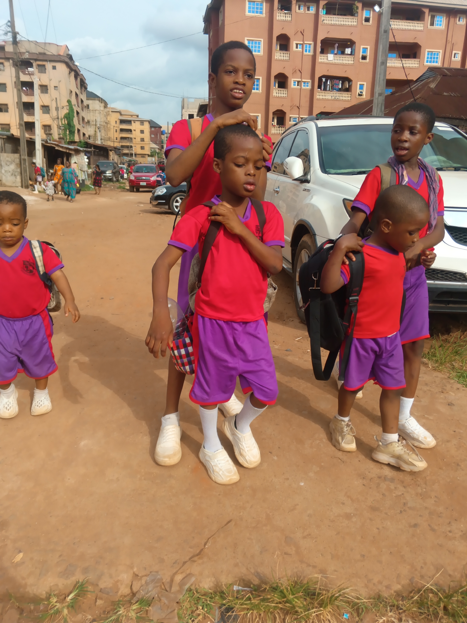 The image shows four children in red shirts and purple shorts, seemingly in Onitsha, Nigeria. They are standing in front of a white SUV on a dirt road in what appears to be a residential area of the city. The children are wearing backpacks and white socks with various types of shoes. Buildings are visible in the background, along with other cars and people in the distance. The sky is overcast. The children appear to be in a school uniform. One of the older children is touching the head of one of the younger children. Another child is holding the backpack of another child.